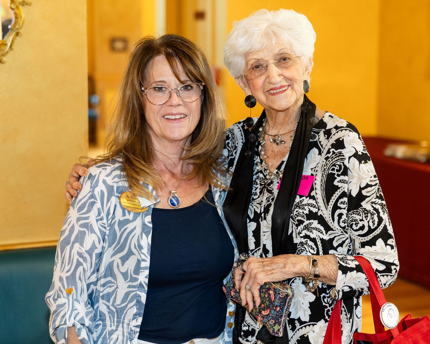 Two smiling women posing together at an indoor event, one in a floral jacket and one in a black floral dress.
