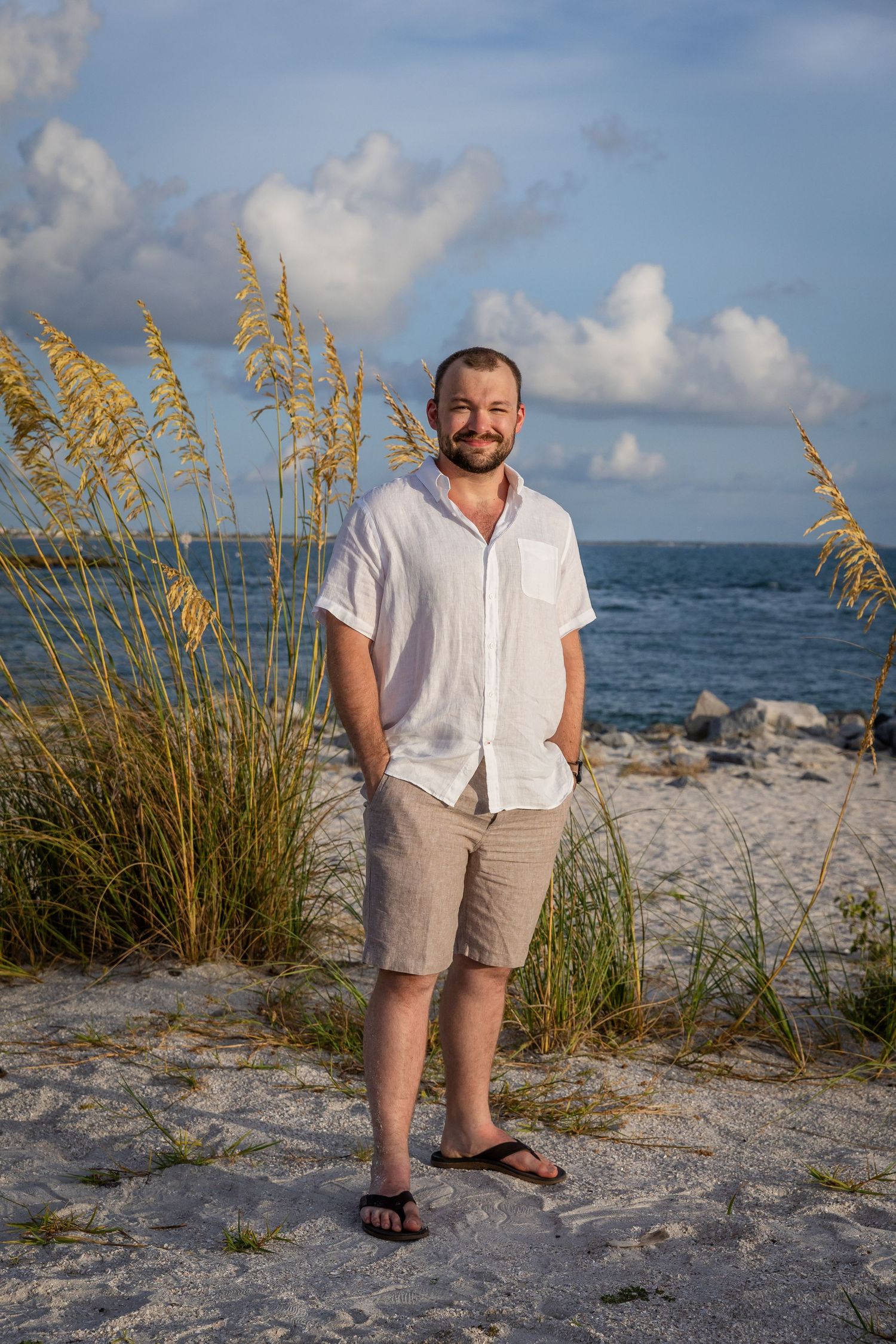 Man in white shirt and khaki shorts smiling on sandy beach with sea oats and blue ocean in background.