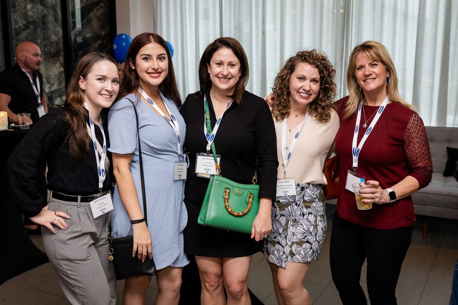 Five women smiling together at a professional networking event, wearing lanyards and name badges indoors.