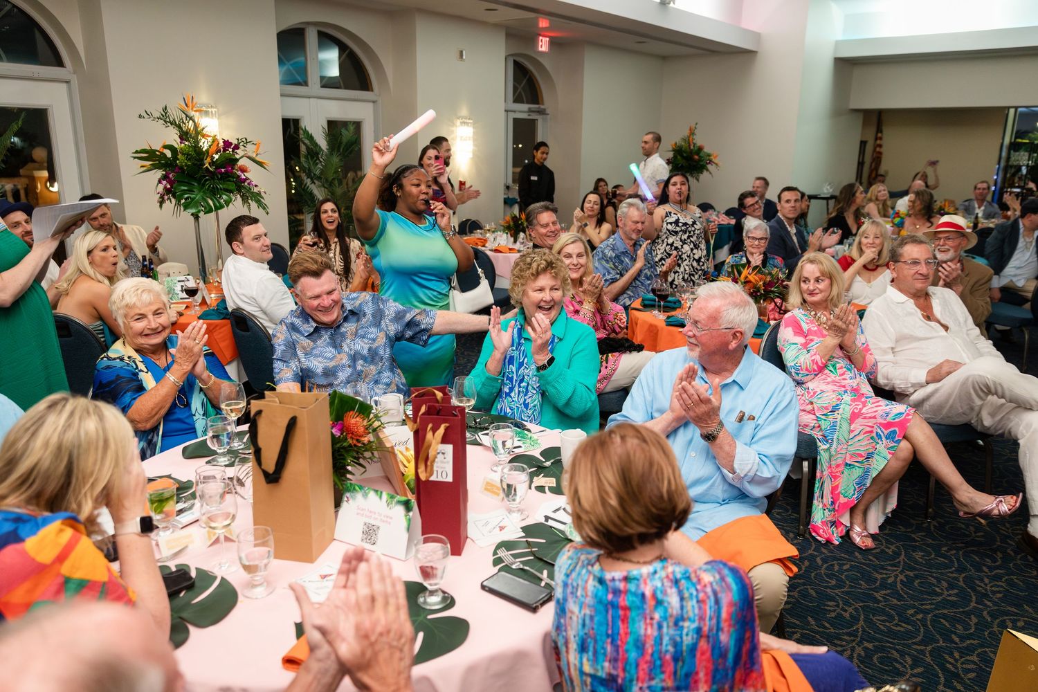 Guests celebrate at a festive indoor party, clapping and cheering around decorated tables with flowers and wine.