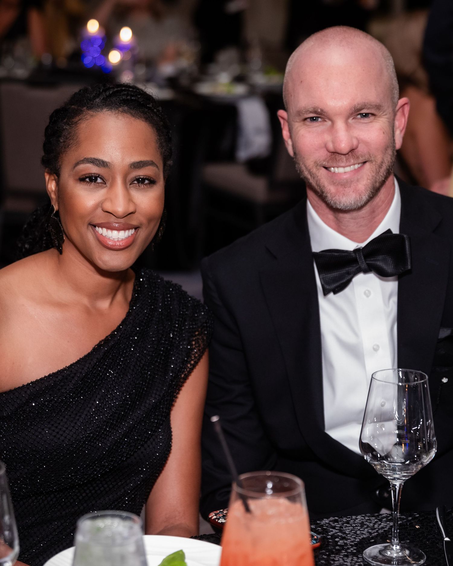 Couple in formal attire smiling at gala dinner table, woman in black dress, man in tuxedo with bow tie.