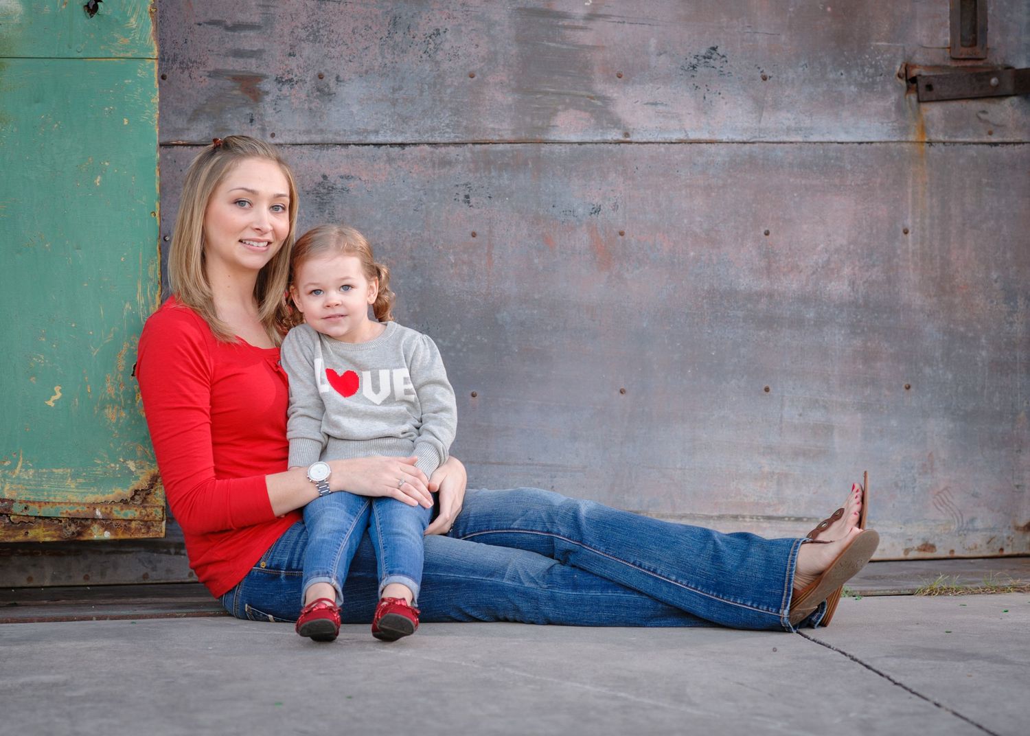 A smiling mother in a red top and jeans sits with her young daughter in a heart sweater against a rustic wall.