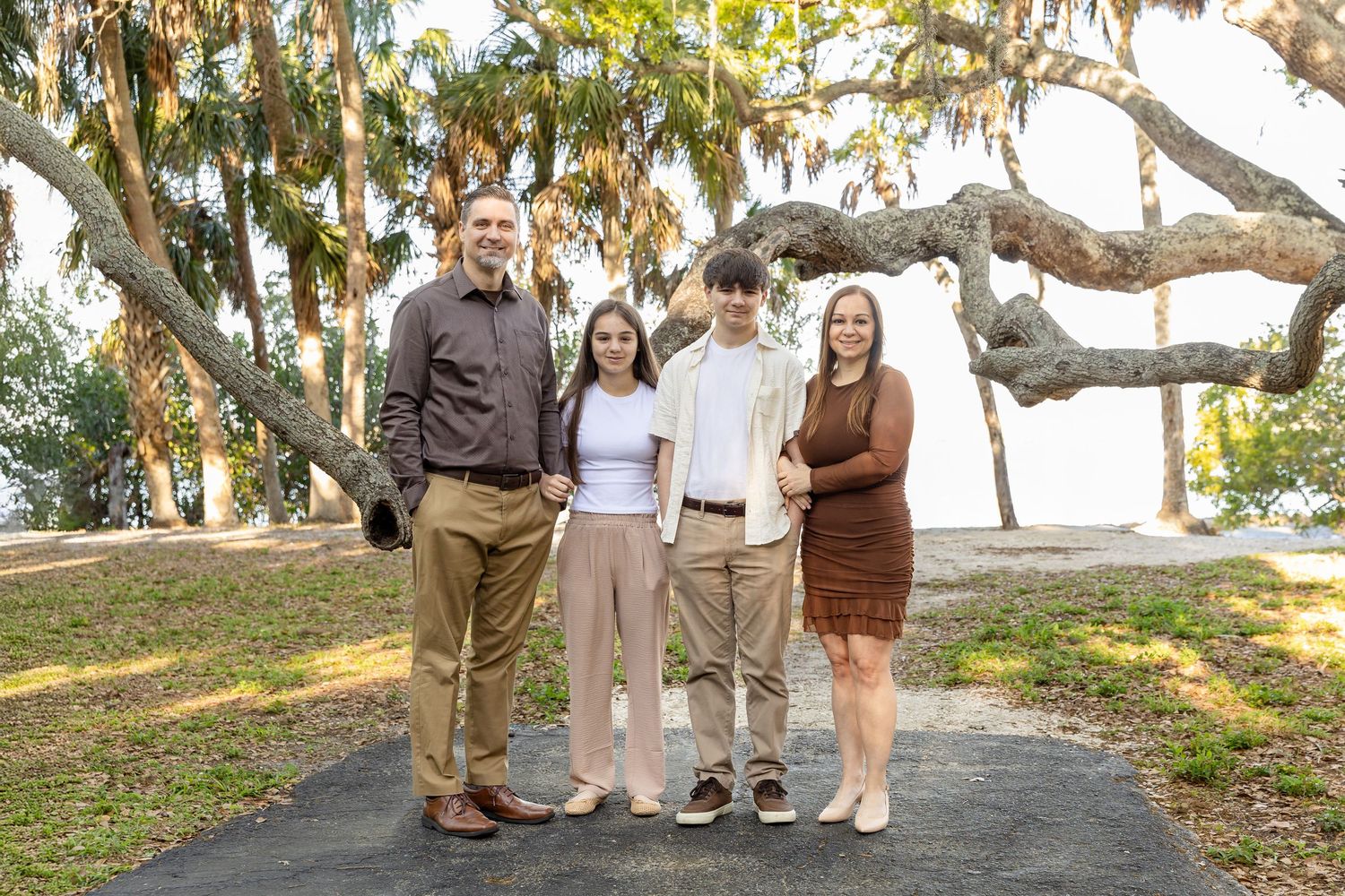 Family of four smiling outdoors under large oak trees on a sunny day in a scenic park setting.