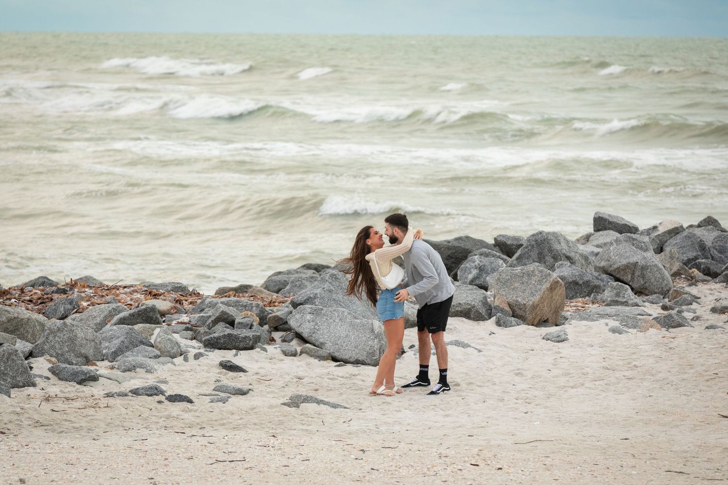 Couple sharing a romantic kiss on a rocky beach with waves crashing in the background.