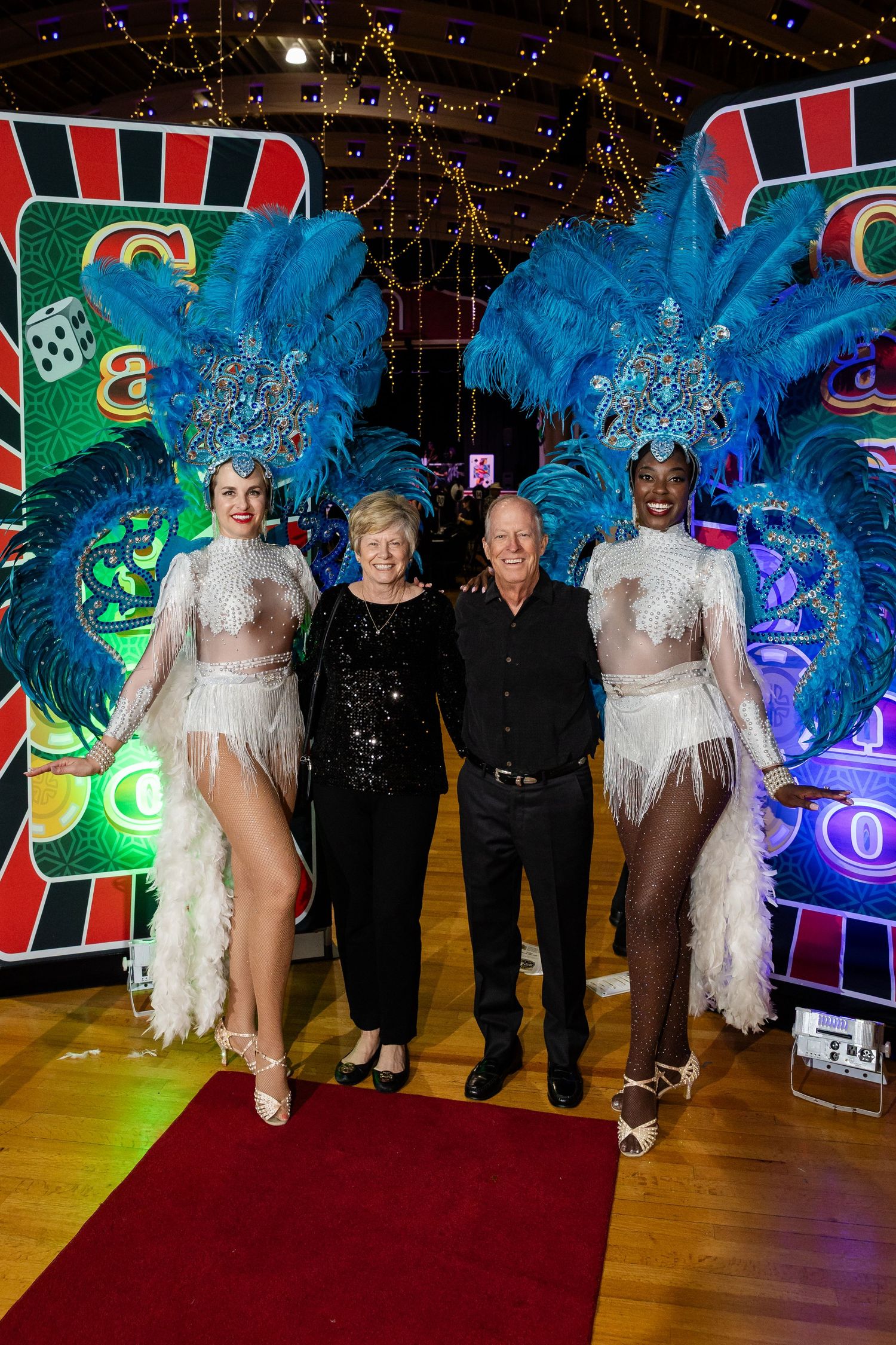 Two showgirls in blue feathered costumes pose with a couple on a red carpet at a casino-themed event.