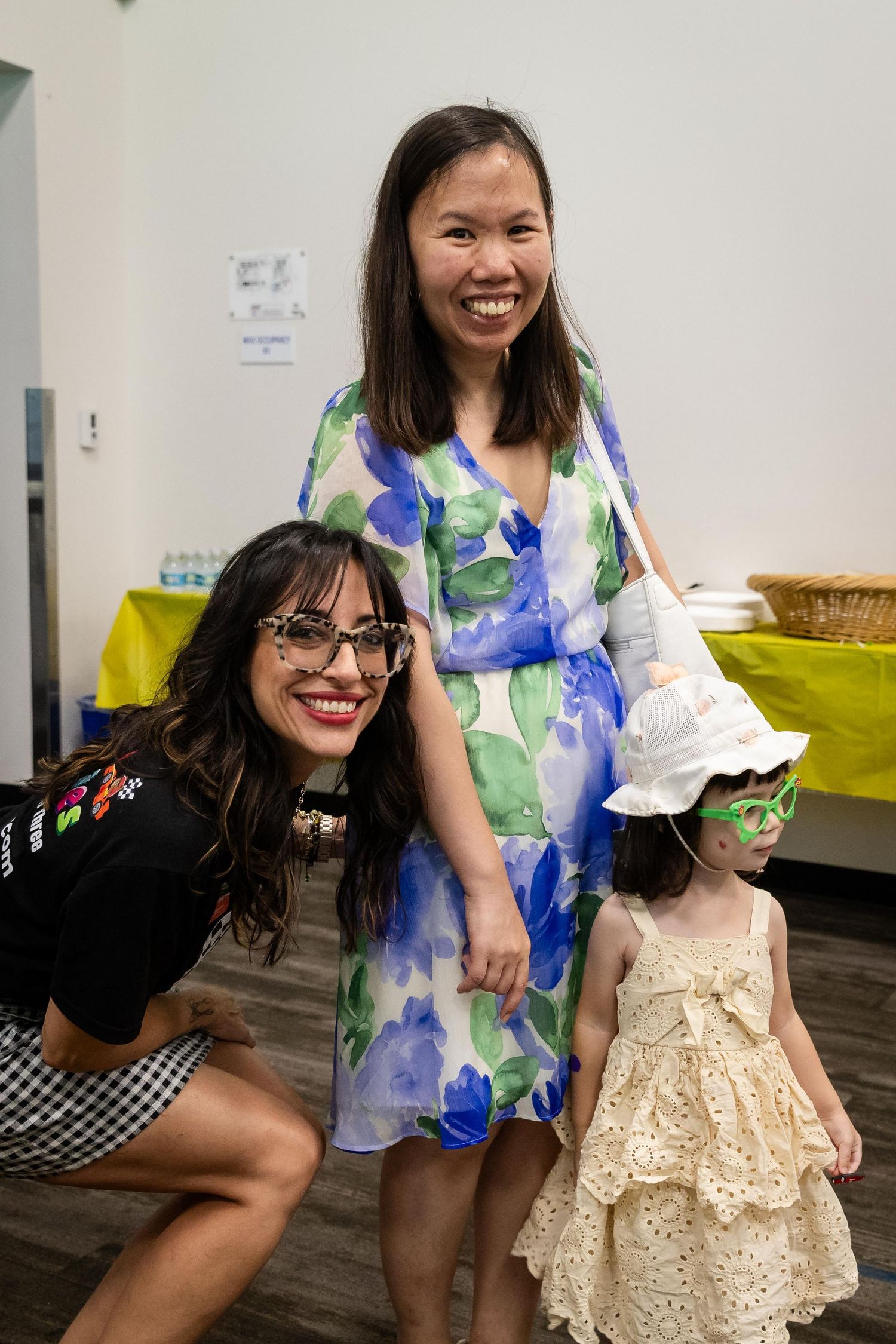 Two women and a young girl in a hat pose together smiling at an indoor event, wearing colorful outfits.