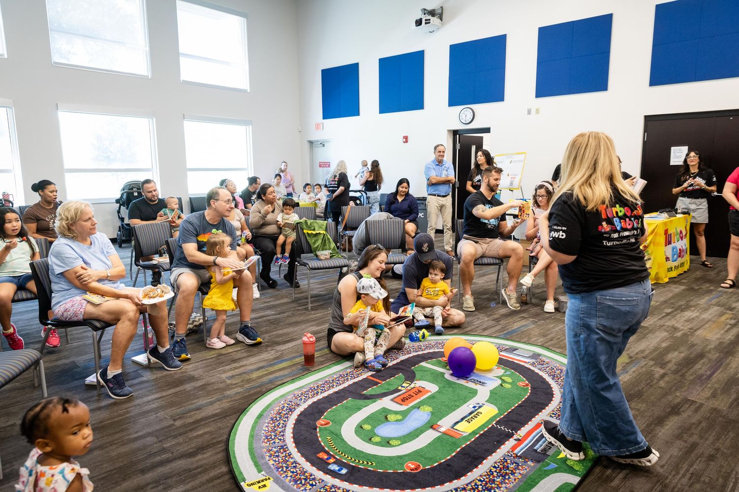 Parents and children gather for an interactive indoor playgroup session with a teacher leading activities on a colorful play mat.