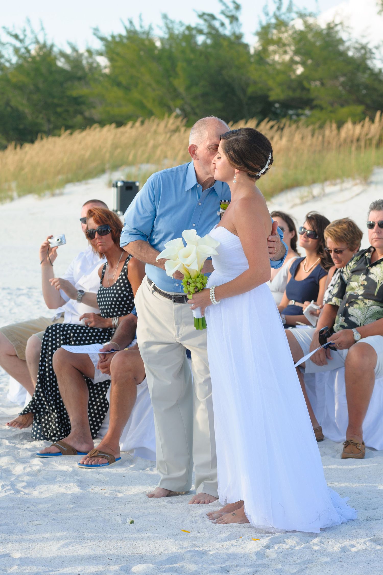 Bride in white strapless gown receives kiss on beach wedding ceremony with seated guests nearby.