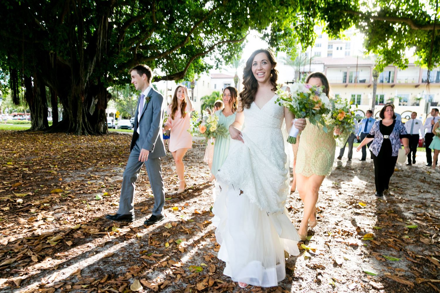 Bride in white gown walks with wedding party under large trees on a leaf-covered path outdoors Museum of Fine Arts, St. Petersburg.