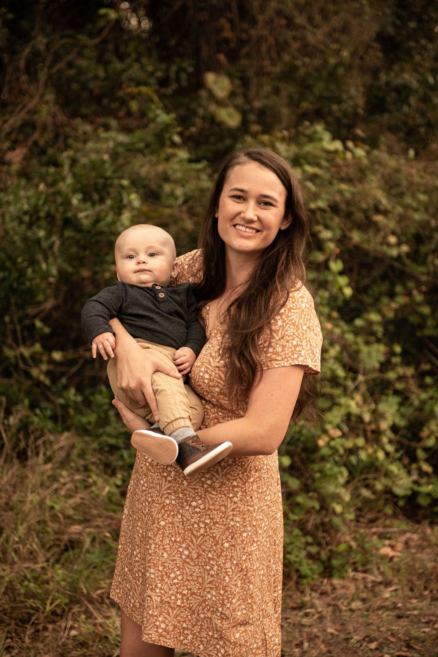 Smiling young woman in floral dress holding a baby in a lush green outdoor setting.