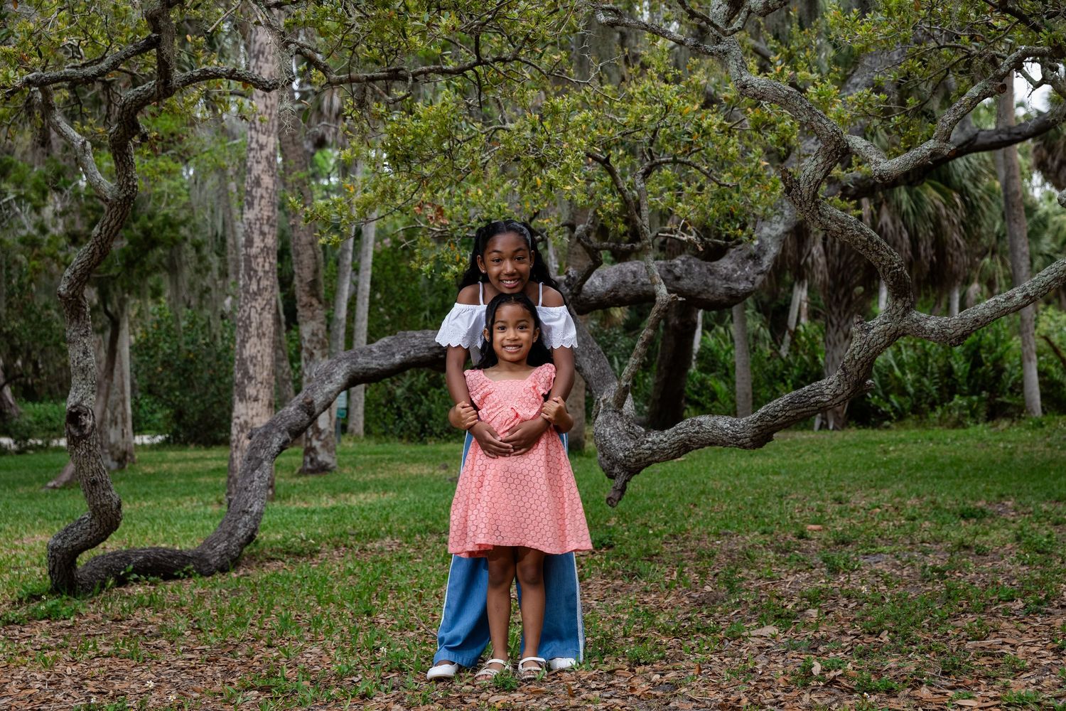 Two girls pose together under a large oak tree in a lush green park, one hugging the other from behind.