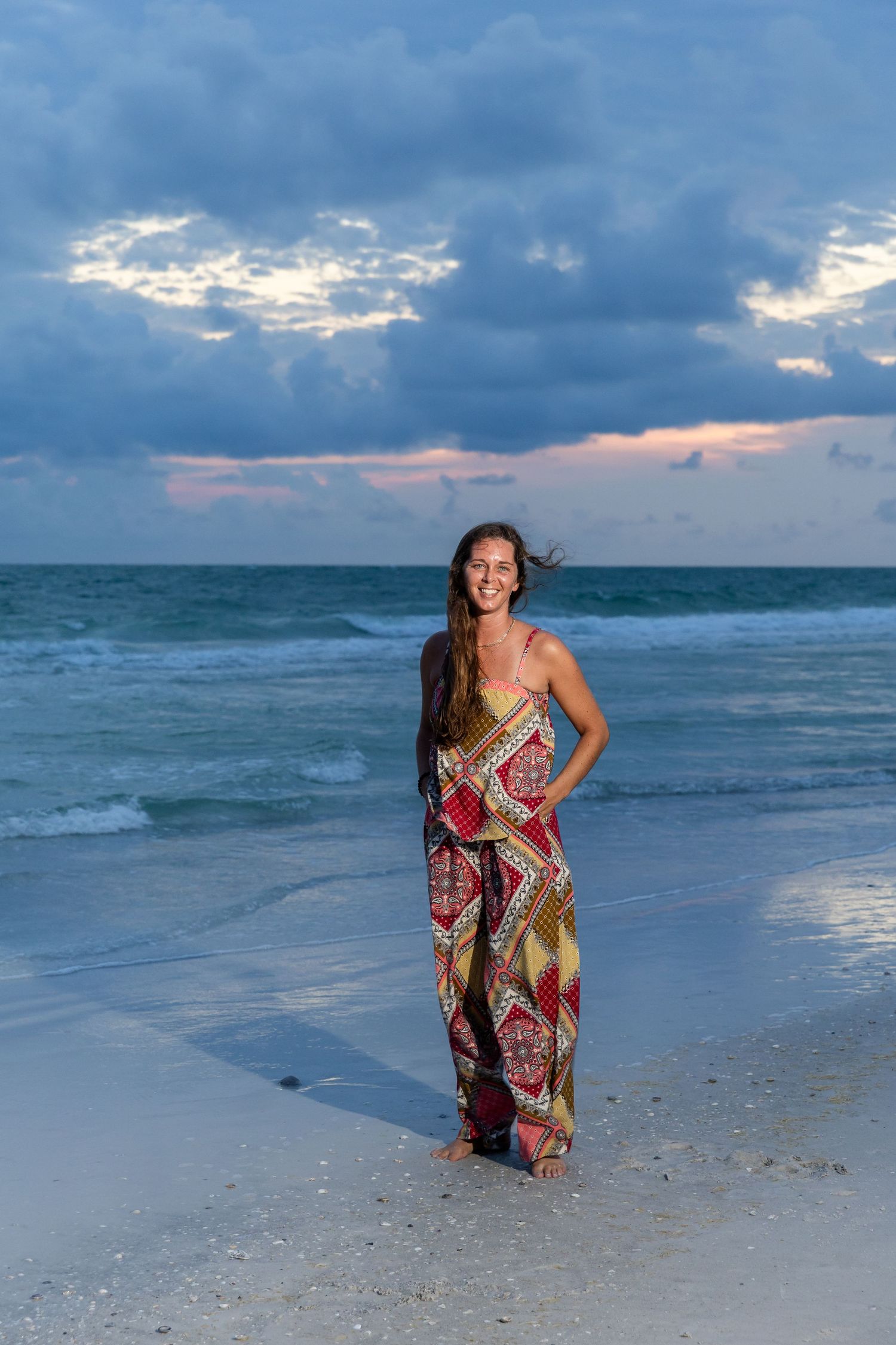 Smiling woman in colorful patterned jumpsuit standing on sandy beach at sunset with dramatic cloudy sky.