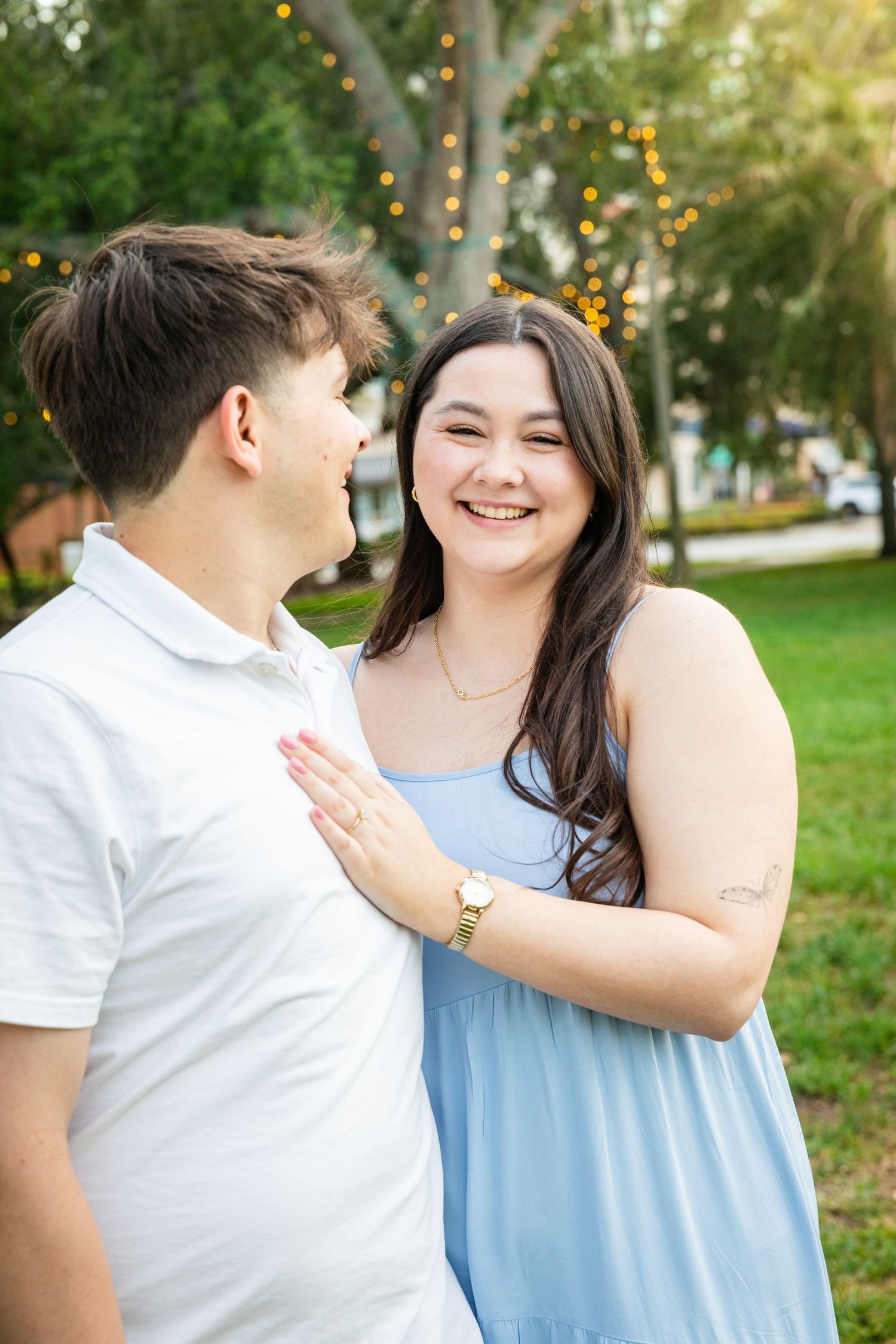 A smiling couple poses outdoors in a park, woman in blue dress with hand on mans chest, string lights in background.