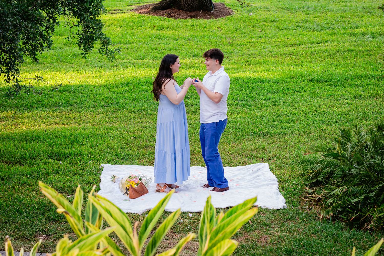 Couple sharing a romantic picnic on a white blanket in a lush green park, woman in blue dress, man in white shirt.