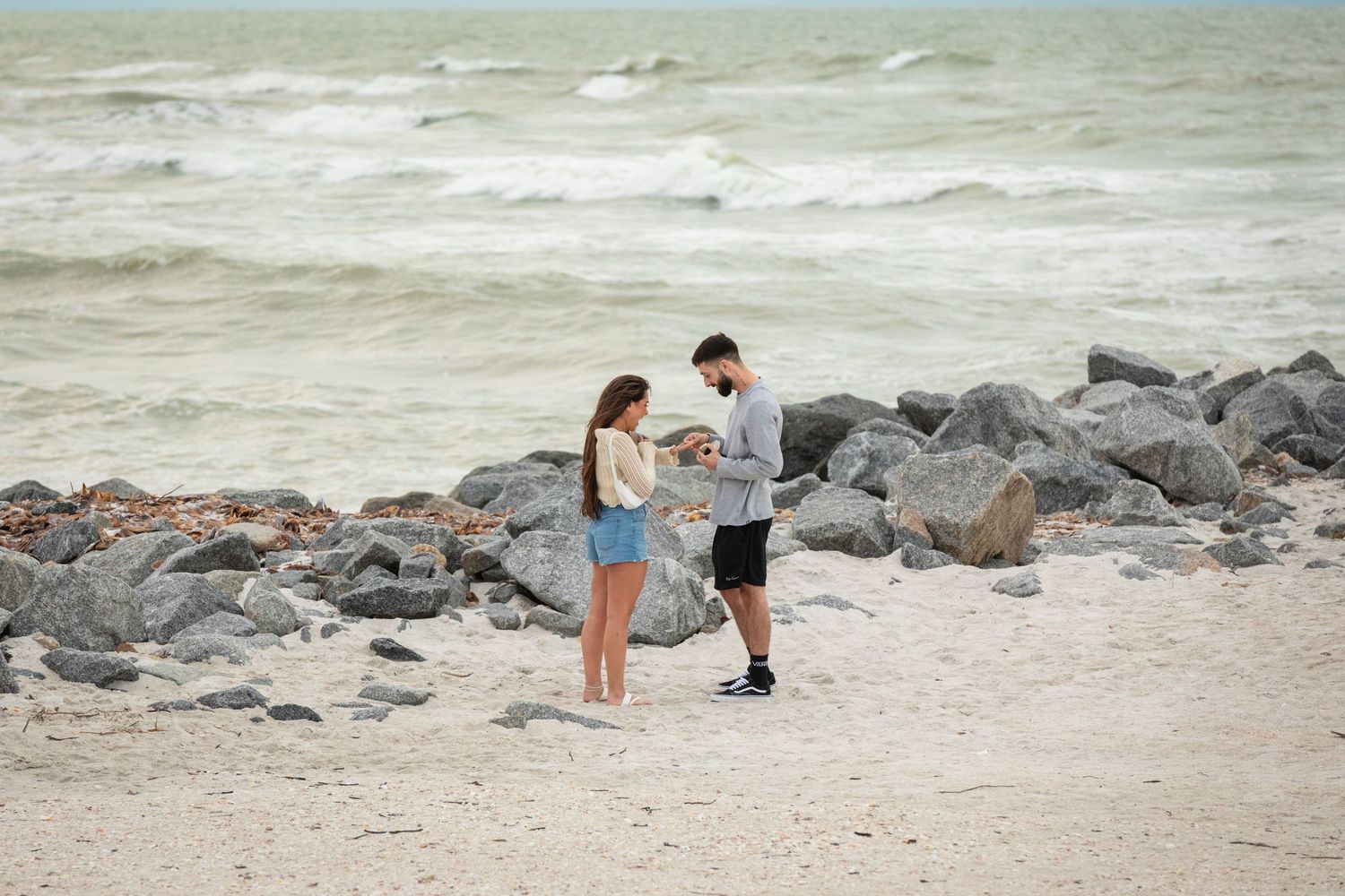 Couple on a sandy beach near rocky shore with turquoise waves, man presenting something to woman.