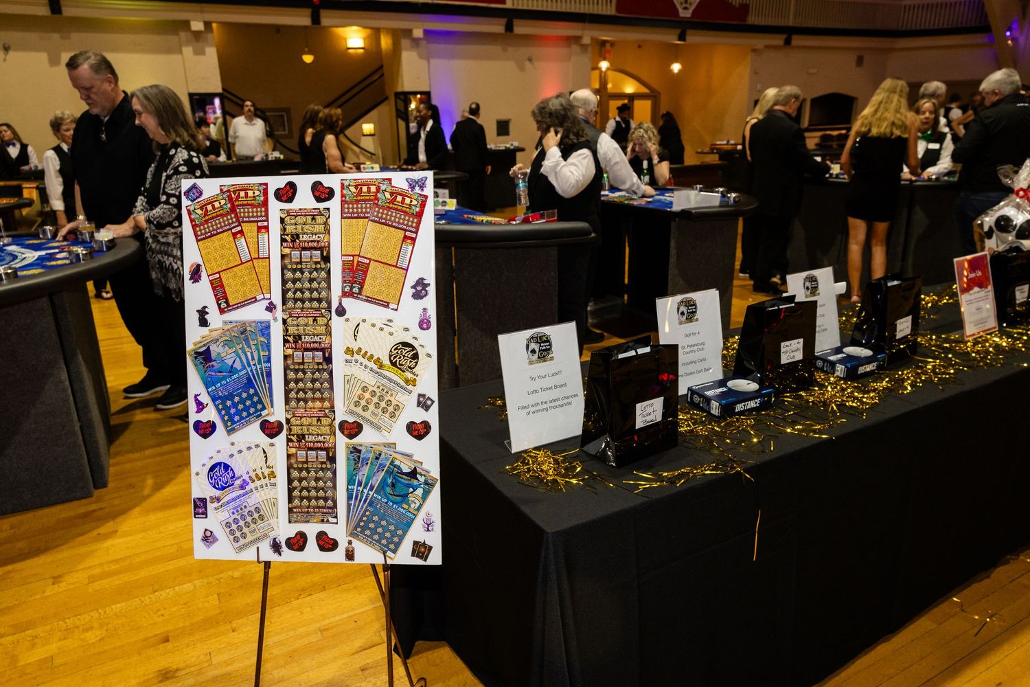 Lottery scratch ticket display board at a charity event with black draped tables and guests mingling in background.