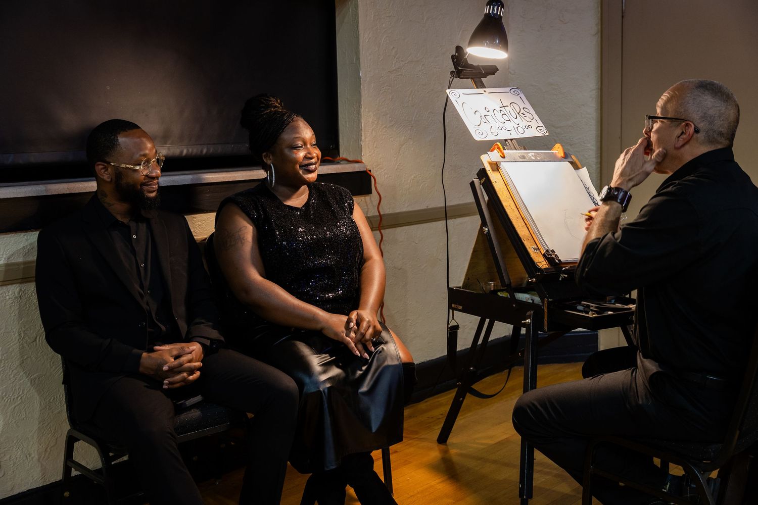 Two people laughing while an artist sketches their portrait at an easel in a dimly lit studio setting.