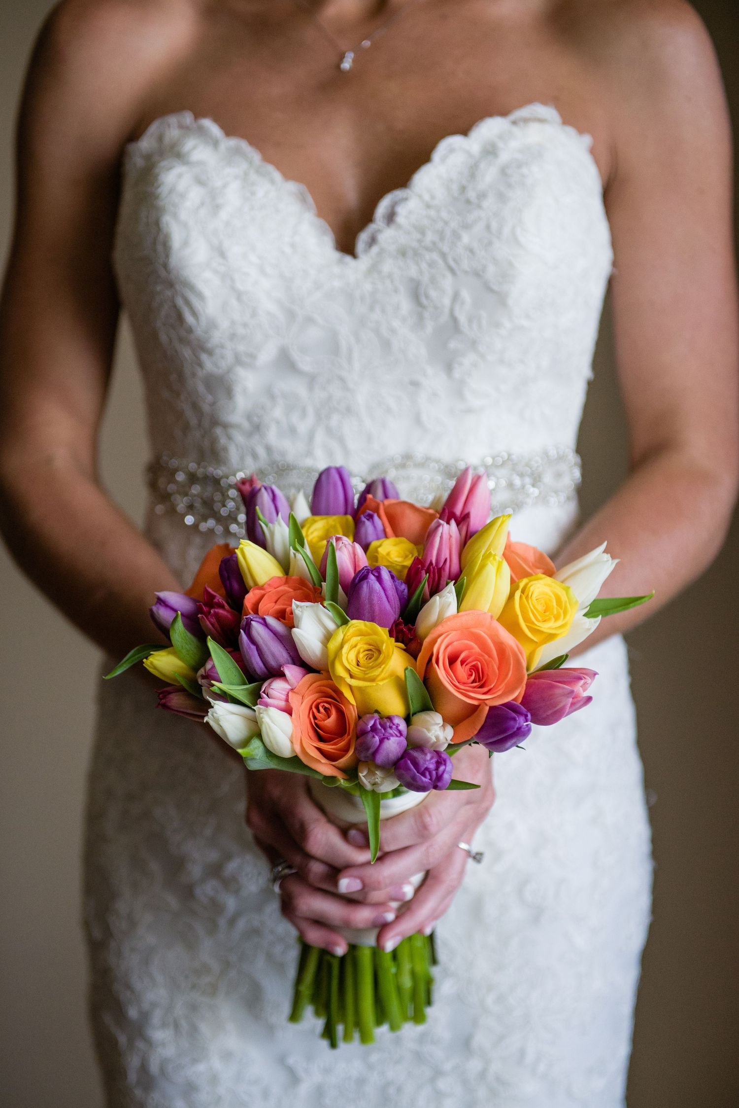Bride in lace strapless gown holding colorful bouquet of roses and tulips.