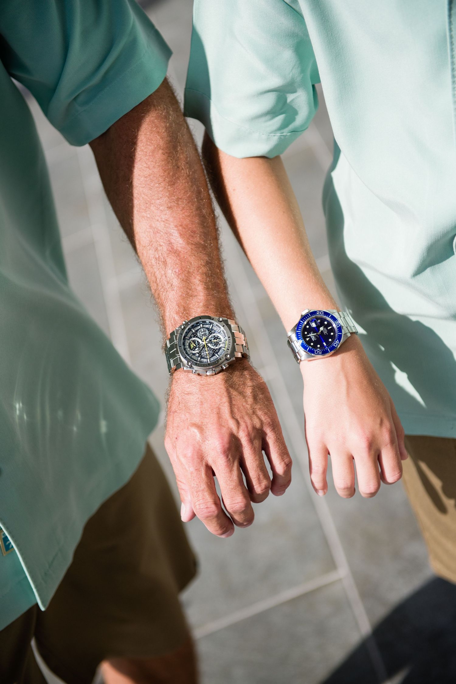 Two people wearing matching luxury watches, one dark chronograph and one blue dial timepiece, with coordinating mint green outfits.