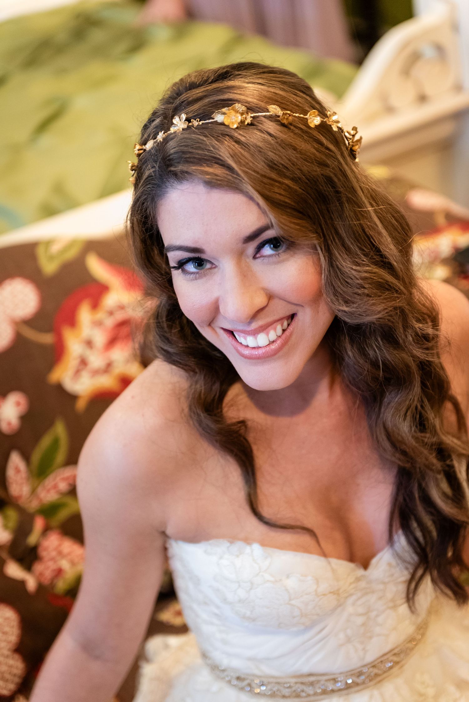 Smiling bride wearing a gold floral crown and strapless lace wedding dress seated on a floral patterned sofa.
