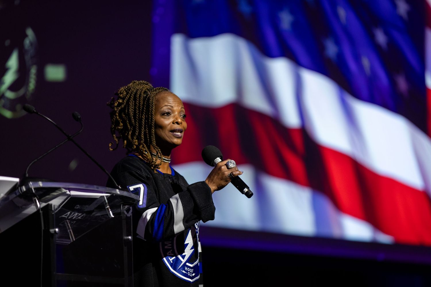 Woman speaker holds microphone at podium with American flag backdrop at formal event.