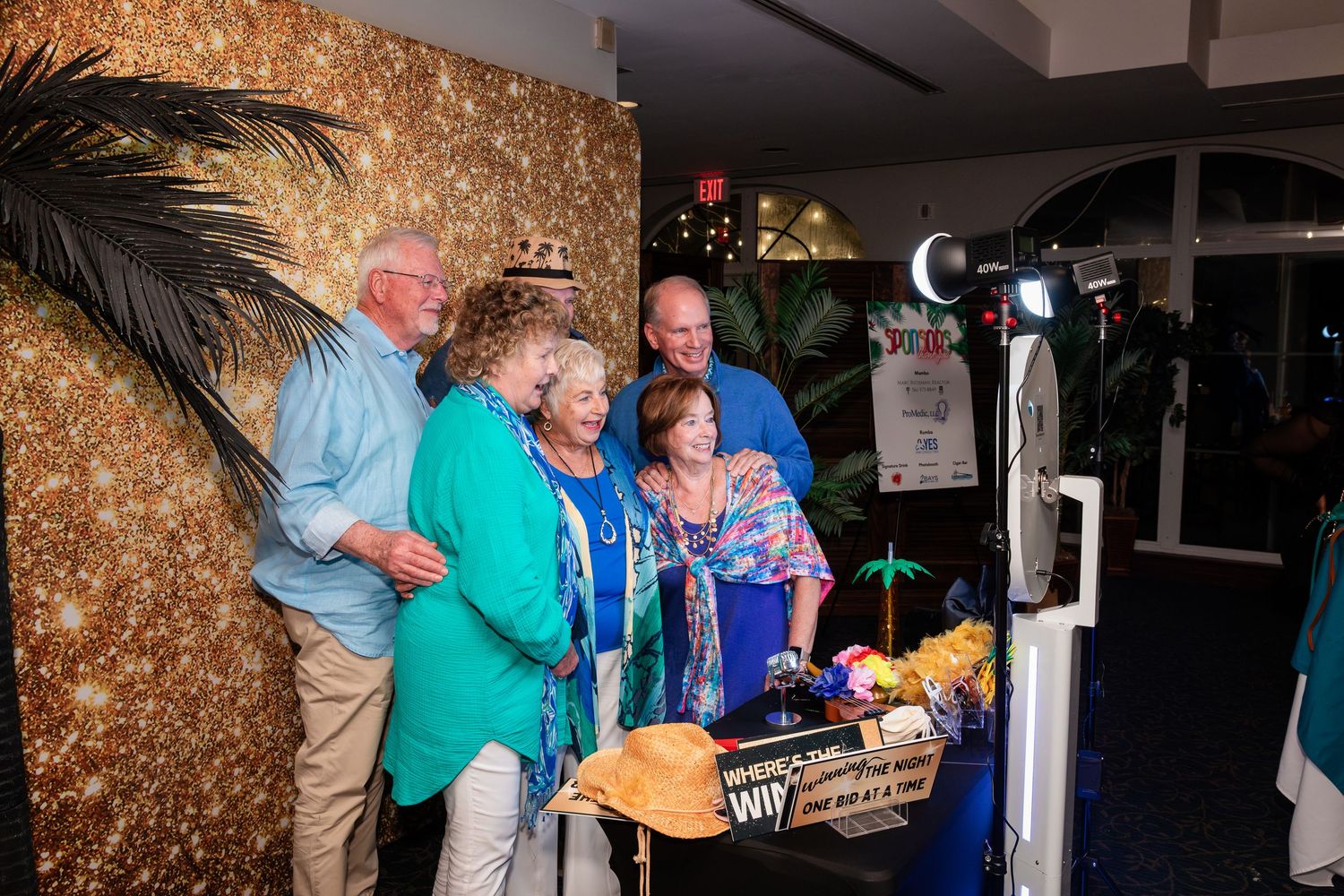 Four adults gather around a photo booth with a gold glitter backdrop at an elegant evening event.