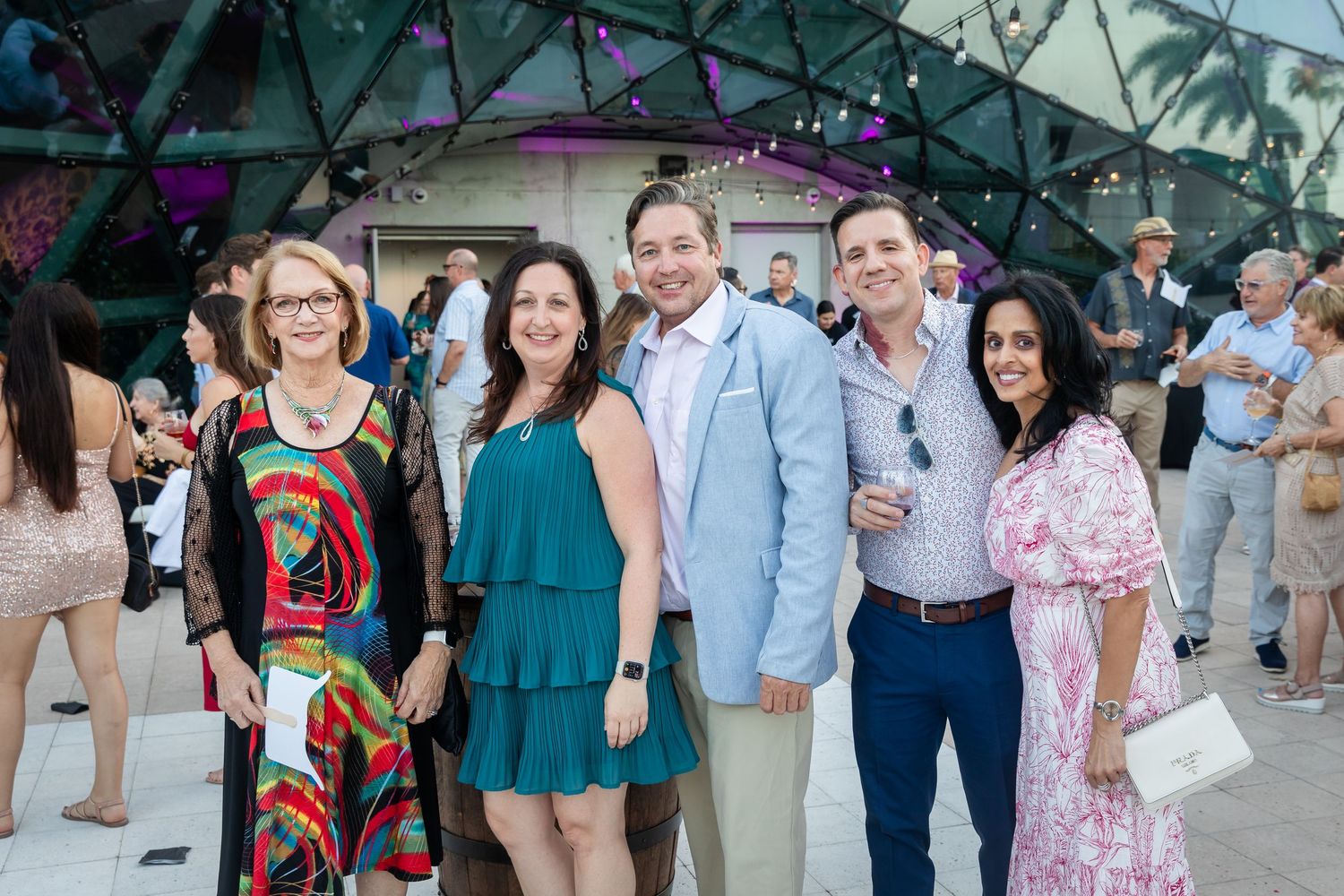 Five friends pose together smiling at an elegant outdoor evening event with colorful lighting.