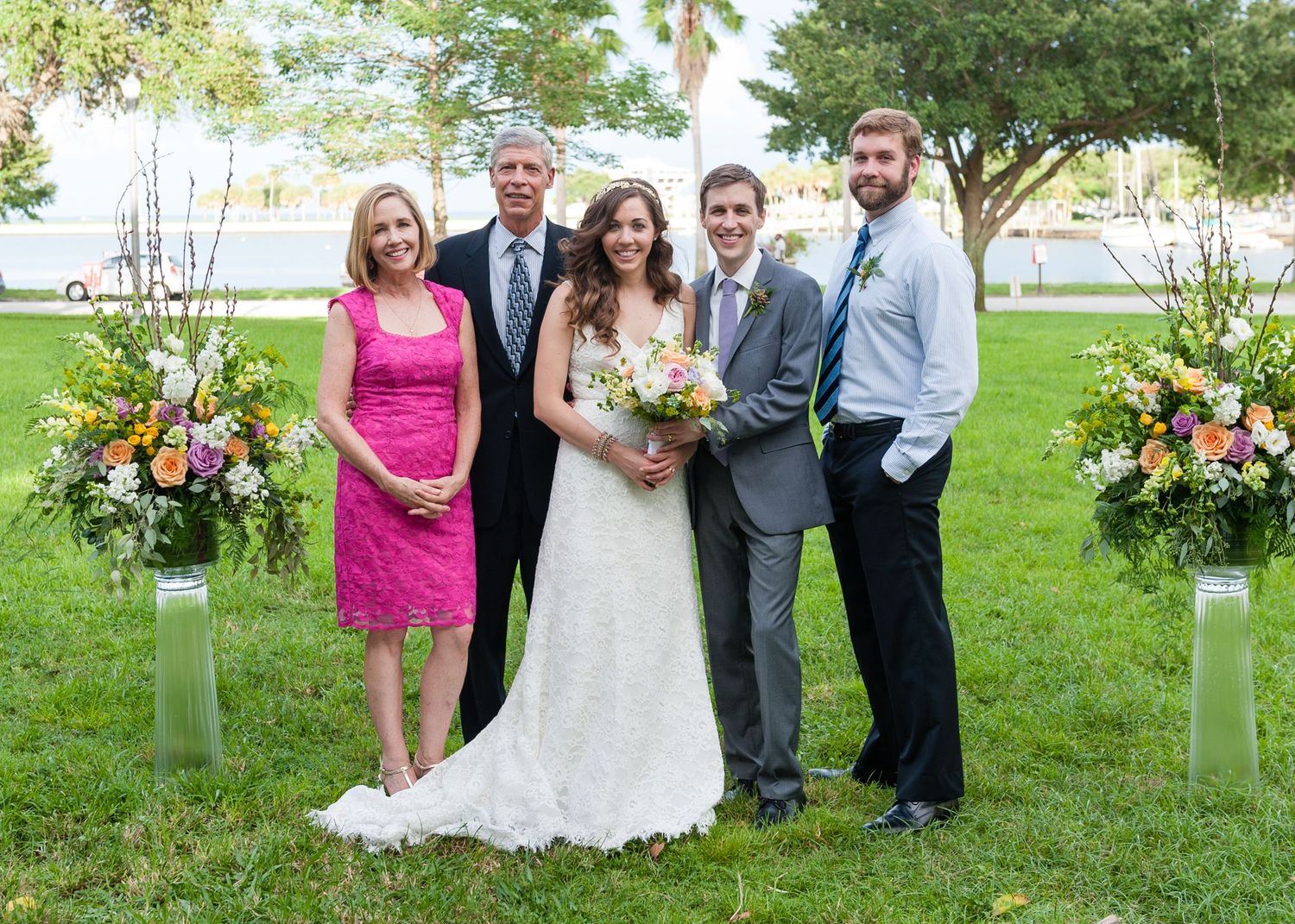 Wedding family portrait with bride and groom flanked by relatives beside colorful floral arrangements outdoors.