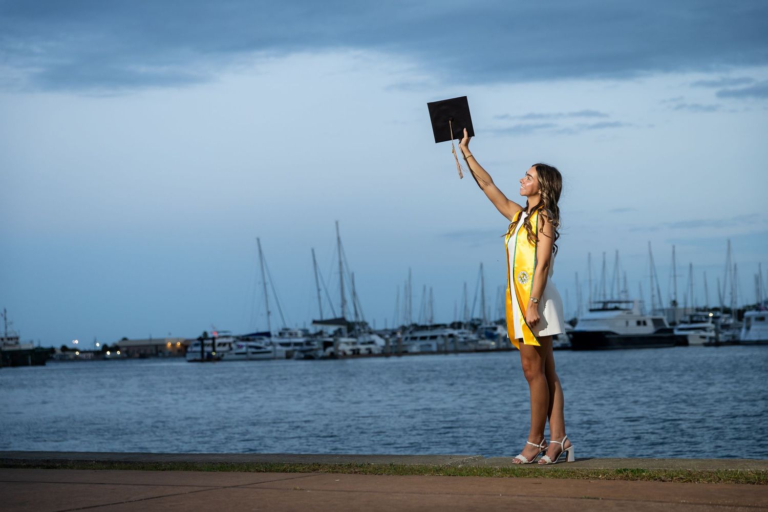 Woman in yellow dress holding tablet aloft near marina at dusk with sailboats in background.