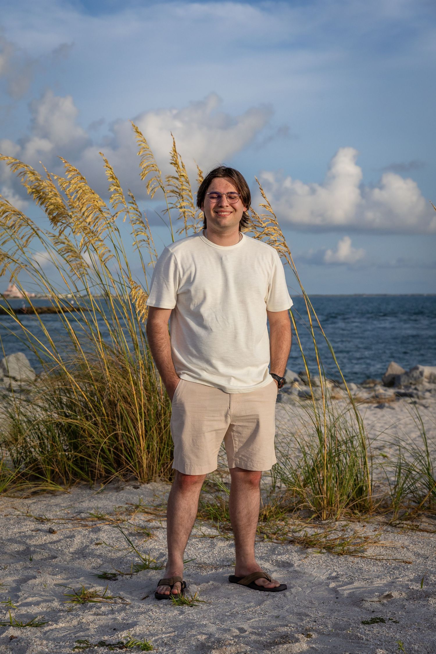 Young man in white tee and khaki shorts smiling on sandy beach with sea oats and ocean backdrop.