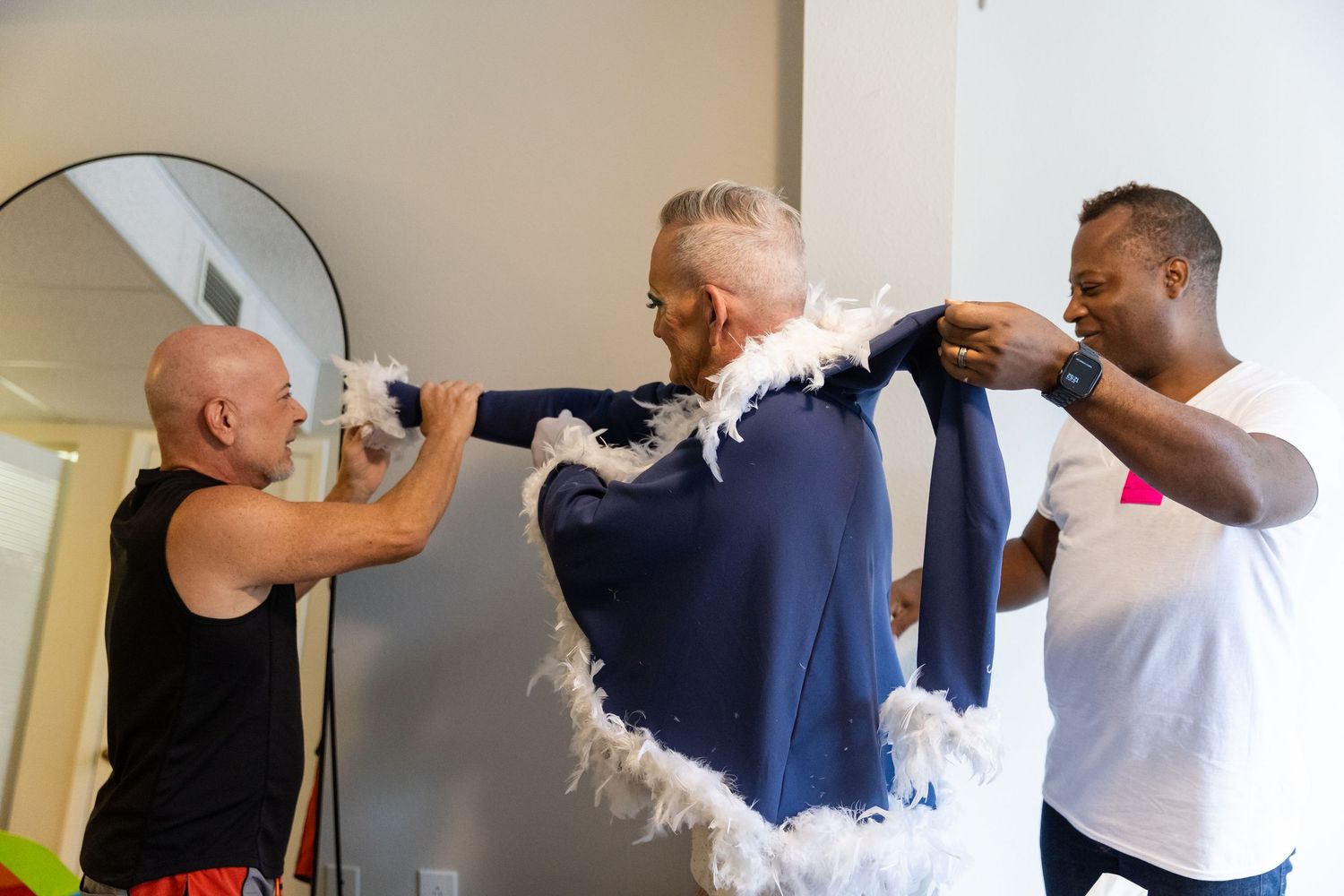 Two men help a third man put on a navy blue jacket with a white feather boa in a bright room.