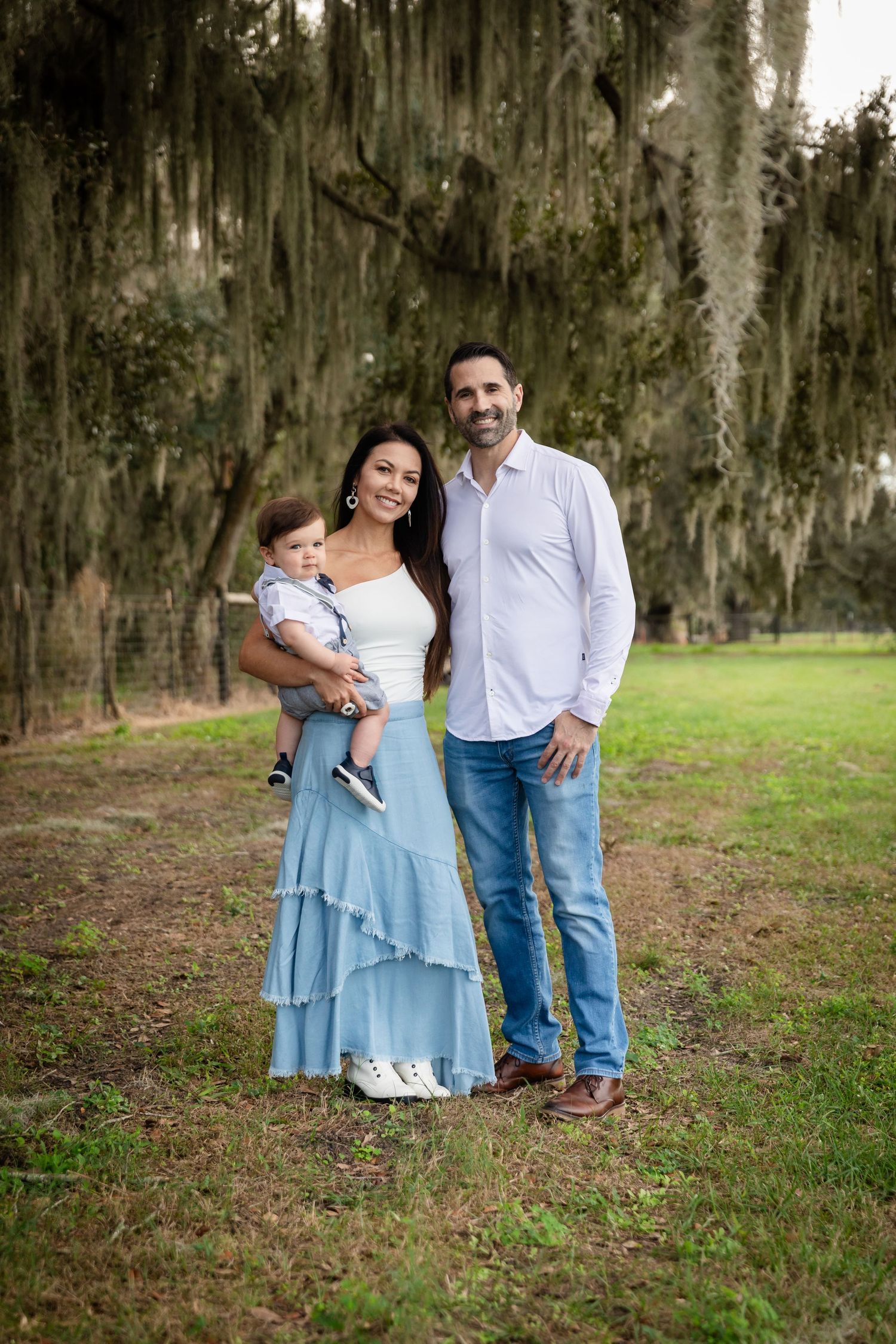 A young family poses outdoors under moss-draped oak trees, mother in blue skirt holding baby, father in white shirt.