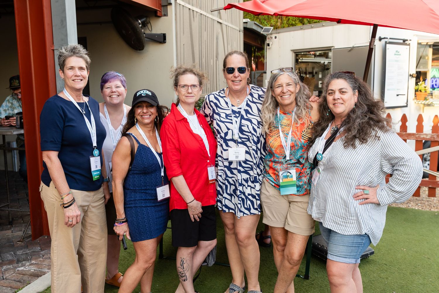 Group of seven women smiling together outdoors at a sunny event, wearing lanyards and casual summer attire.