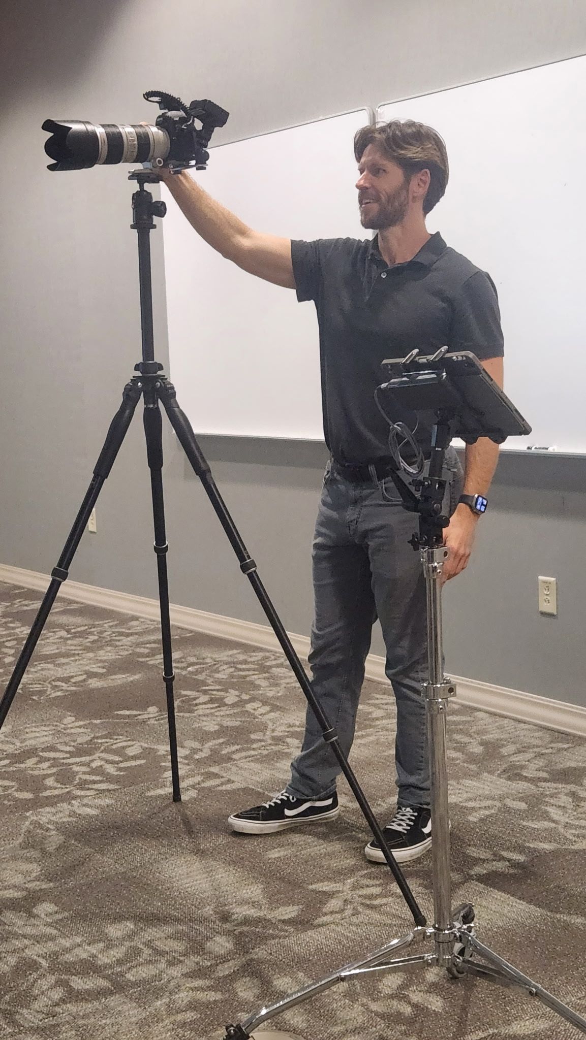 A photographer adjusts a camera with telephoto lens on a tripod in a conference room, wearing a black polo shirt.