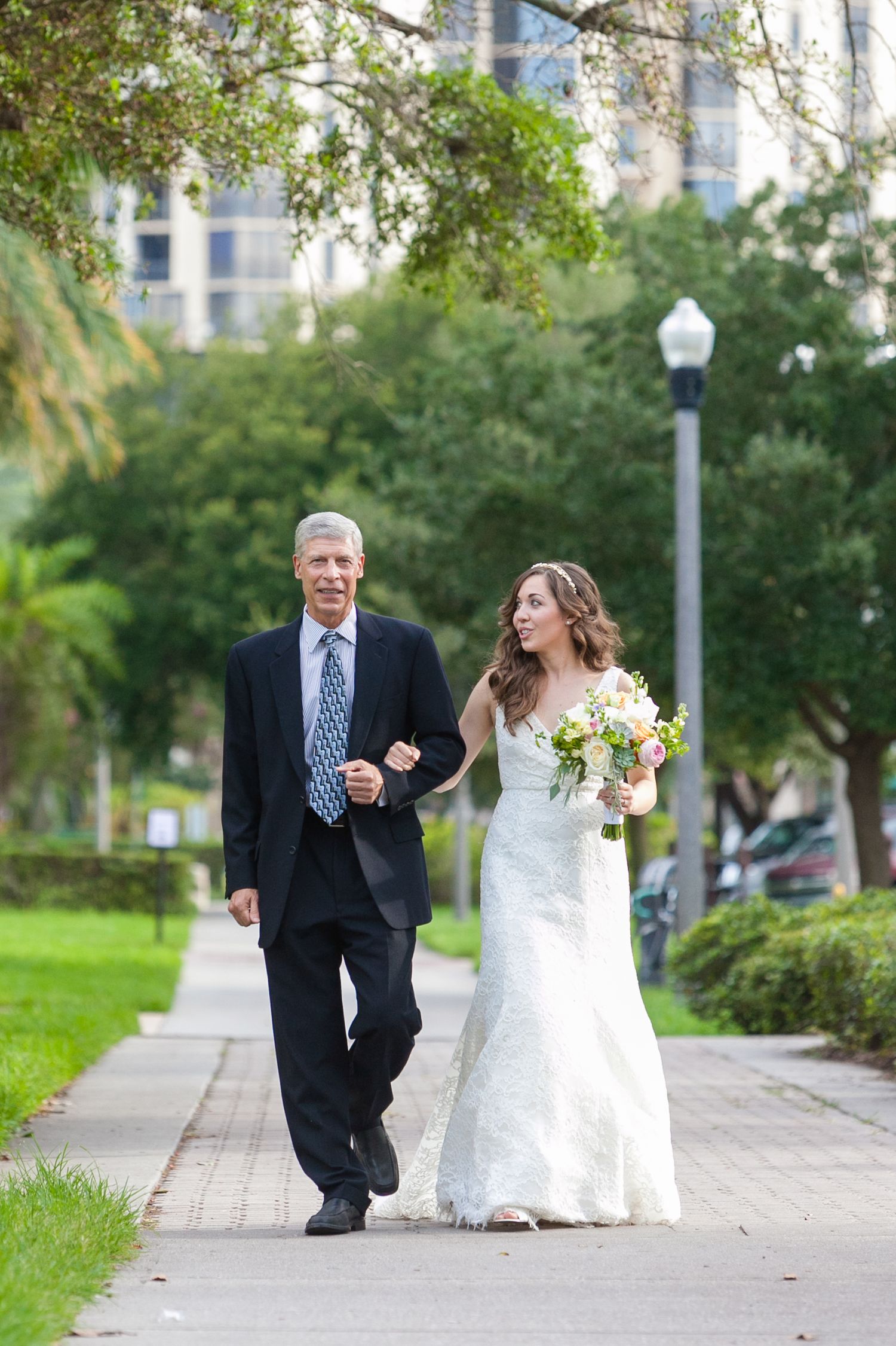 Bride in white lace gown walking arm in arm with father in dark suit on pathway Museum of Fine Arts, St. Petersburg.