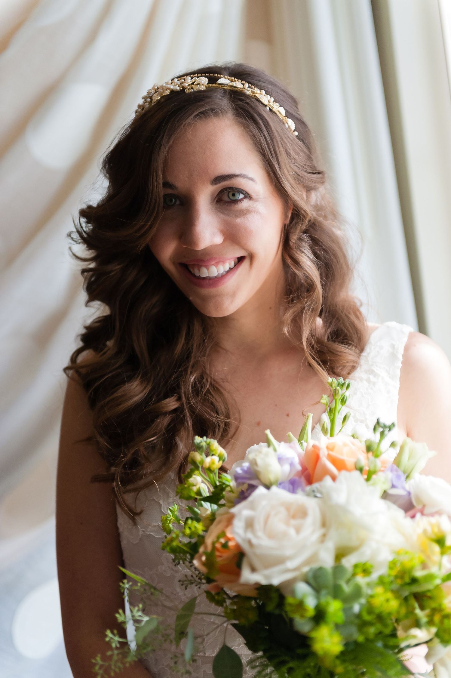 Beautiful bride with curly hair and a gold headpiece smiling while holding a colorful floral bouquet.