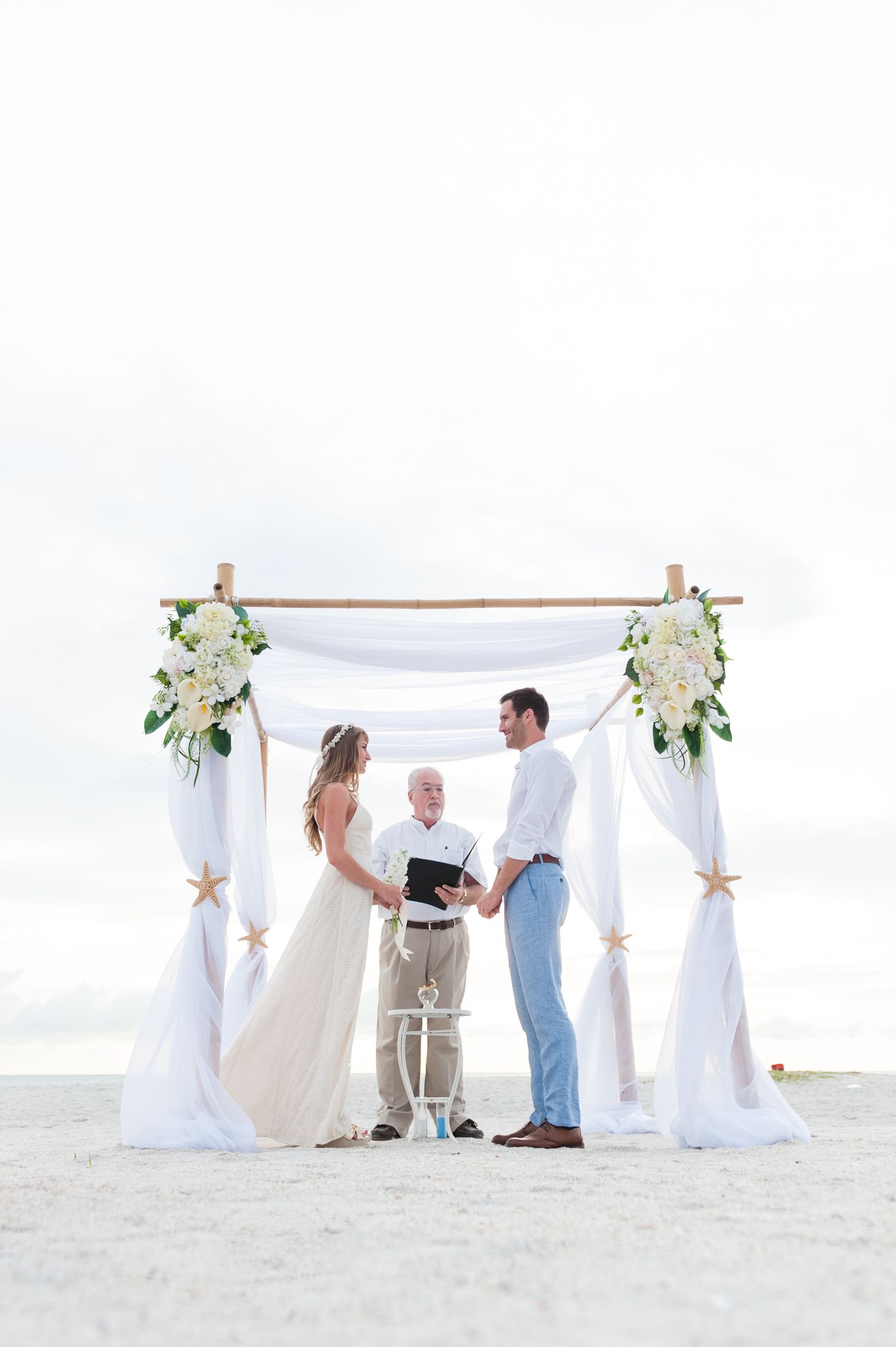 Bride and groom exchange vows under elegant bamboo arch adorned with white flowers on the beach on Treasure Island, Florida.