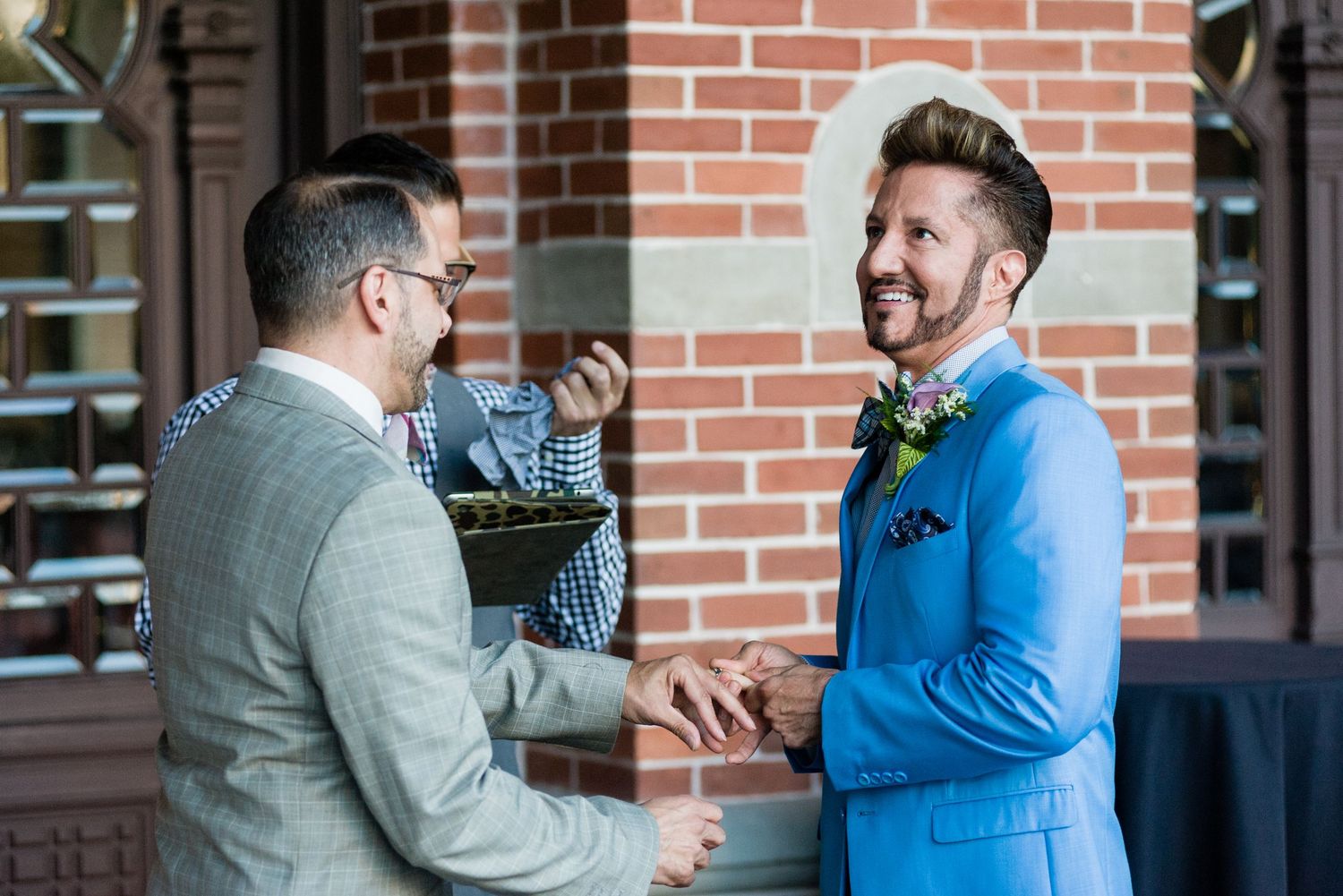 Two grooms in suits smile and hold hands at wedding ceremony.