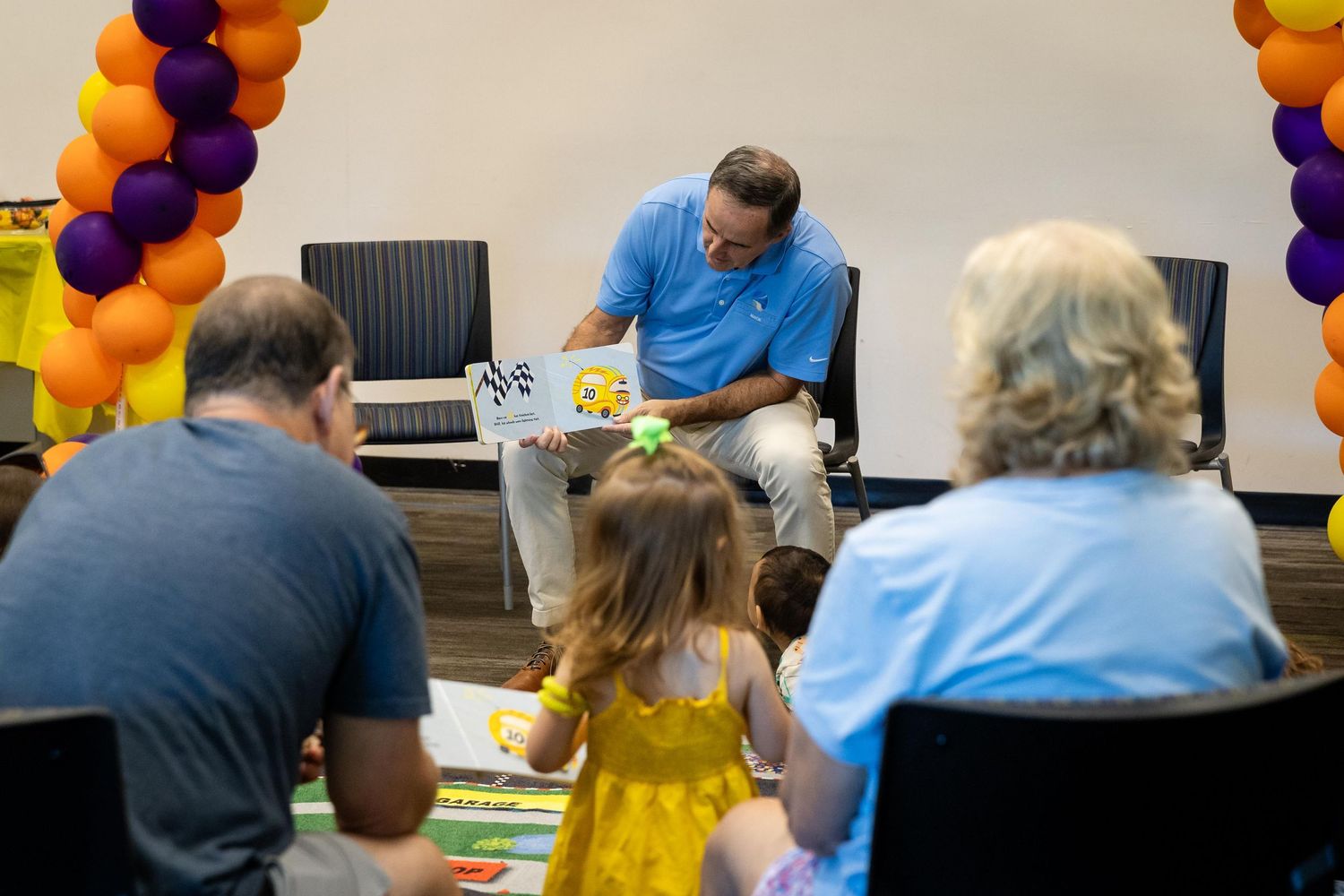 A man in a blue shirt reads a picture book to young children and adults in a colorful balloon-decorated room.