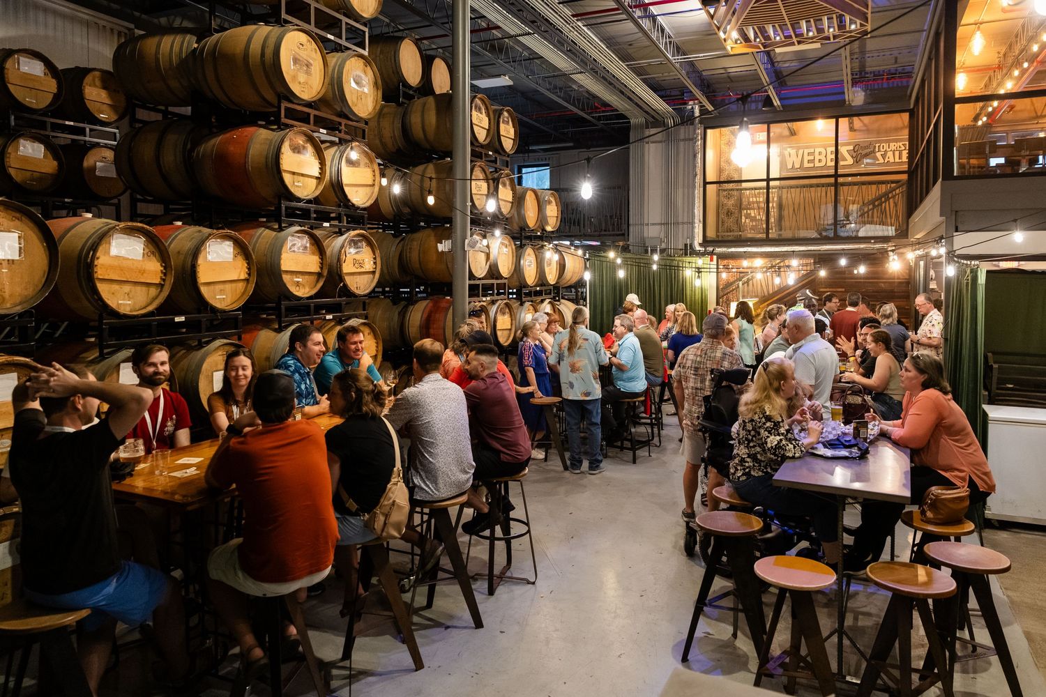 Guests gather at barrel-lined winery tasting room with string lights, enjoying wine at rustic tables in a warm, inviting atmosphere.