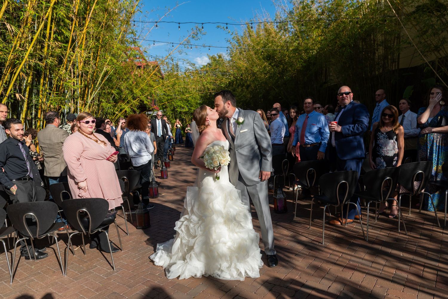 Bride and groom share first kiss at outdoor wedding ceremony lined with bamboo trees and seated guests.