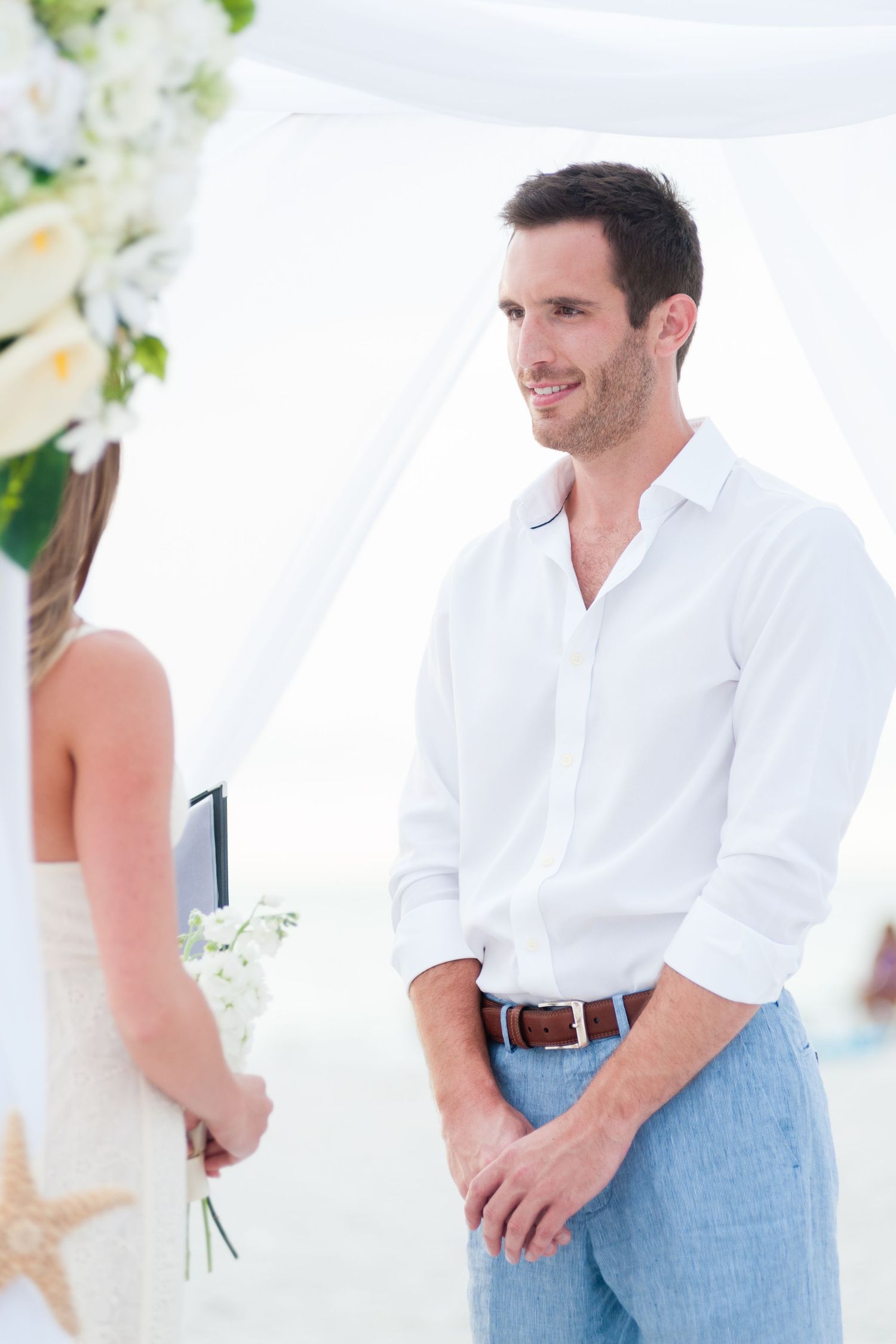 Groom in white shirt and blue jeans smiling at bride during outdoor beach wedding ceremony.