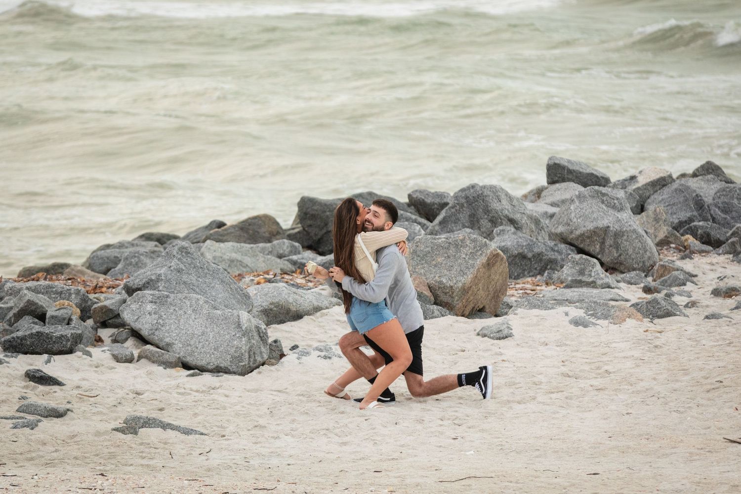 Couple embracing romantically on a sandy beach with rocky shoreline and ocean waves in the background.