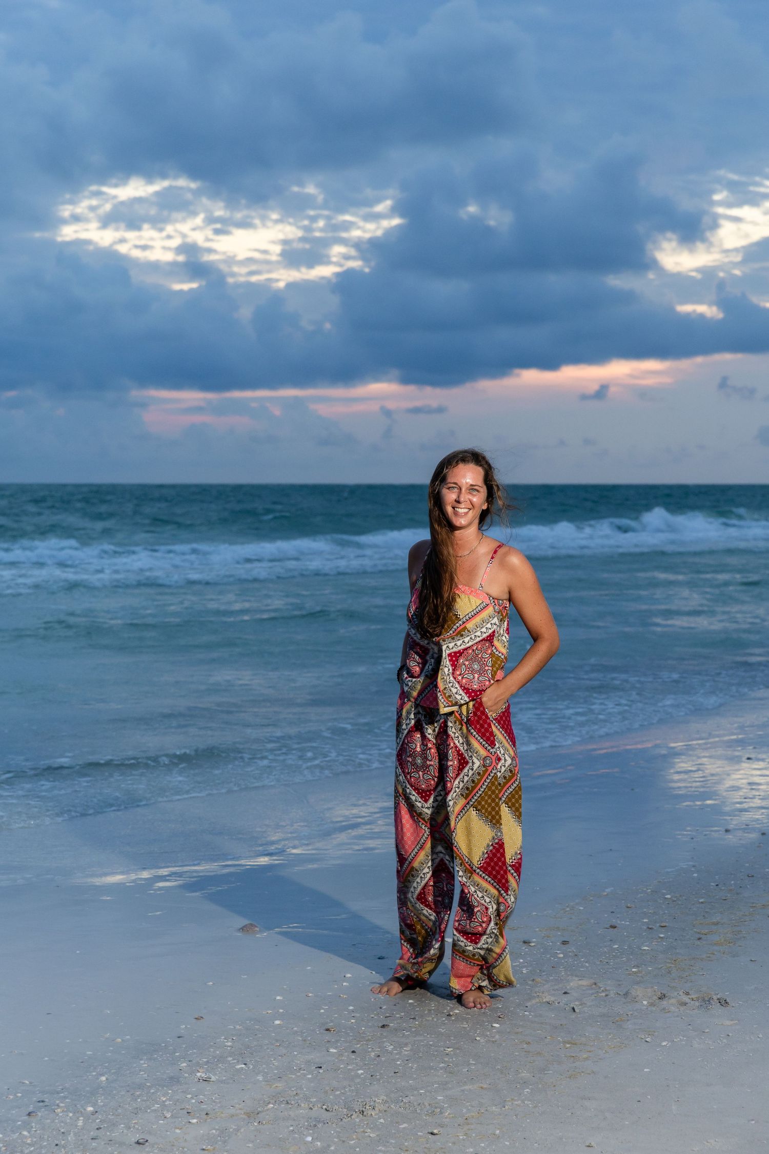 Young woman in colorful maxi dress smiling on sandy beach at sunset with dramatic cloudy sky.