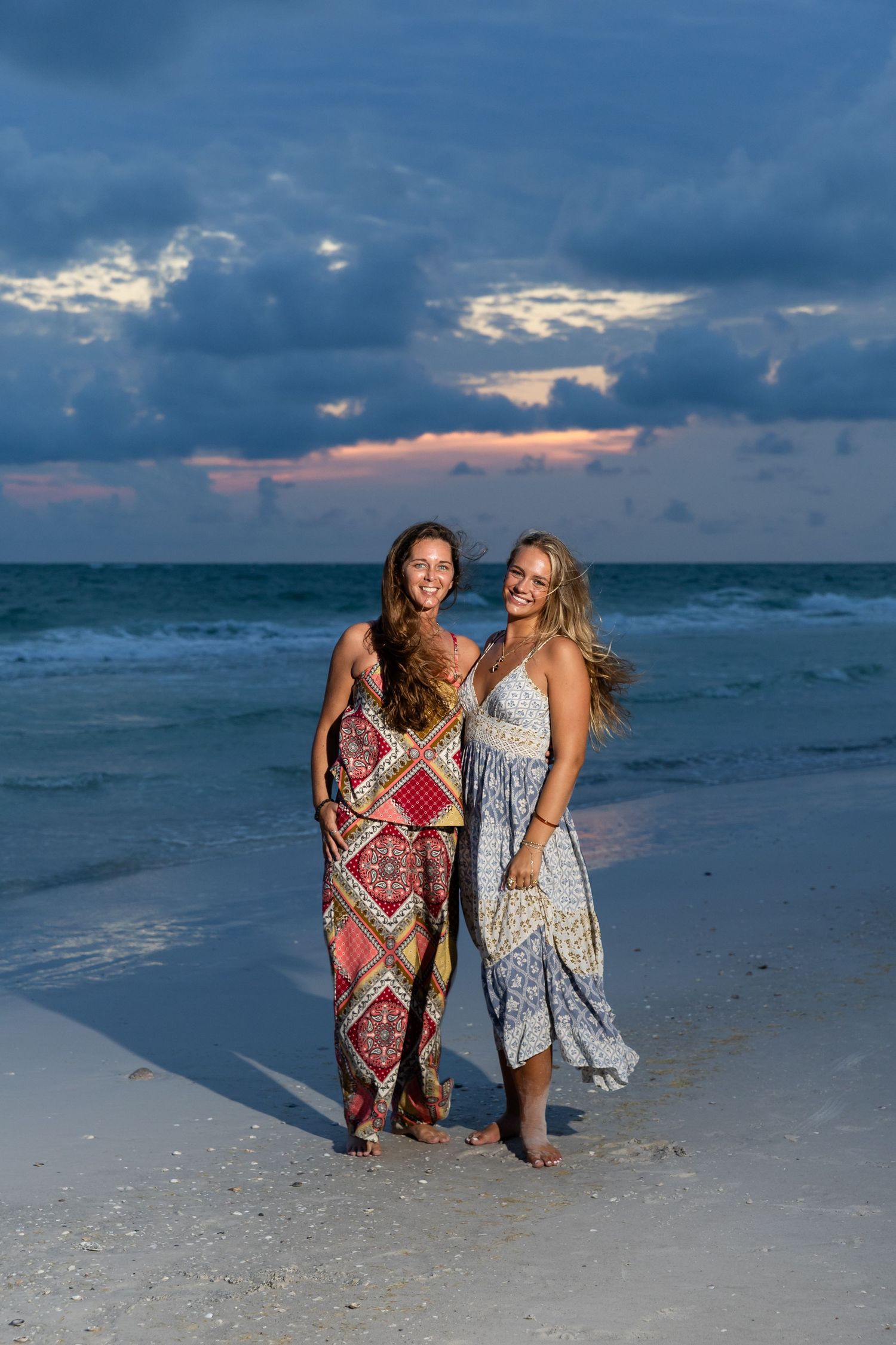 Two women in boho maxi dresses smile on a sandy beach at sunset with dramatic cloudy skies and ocean waves behind them.