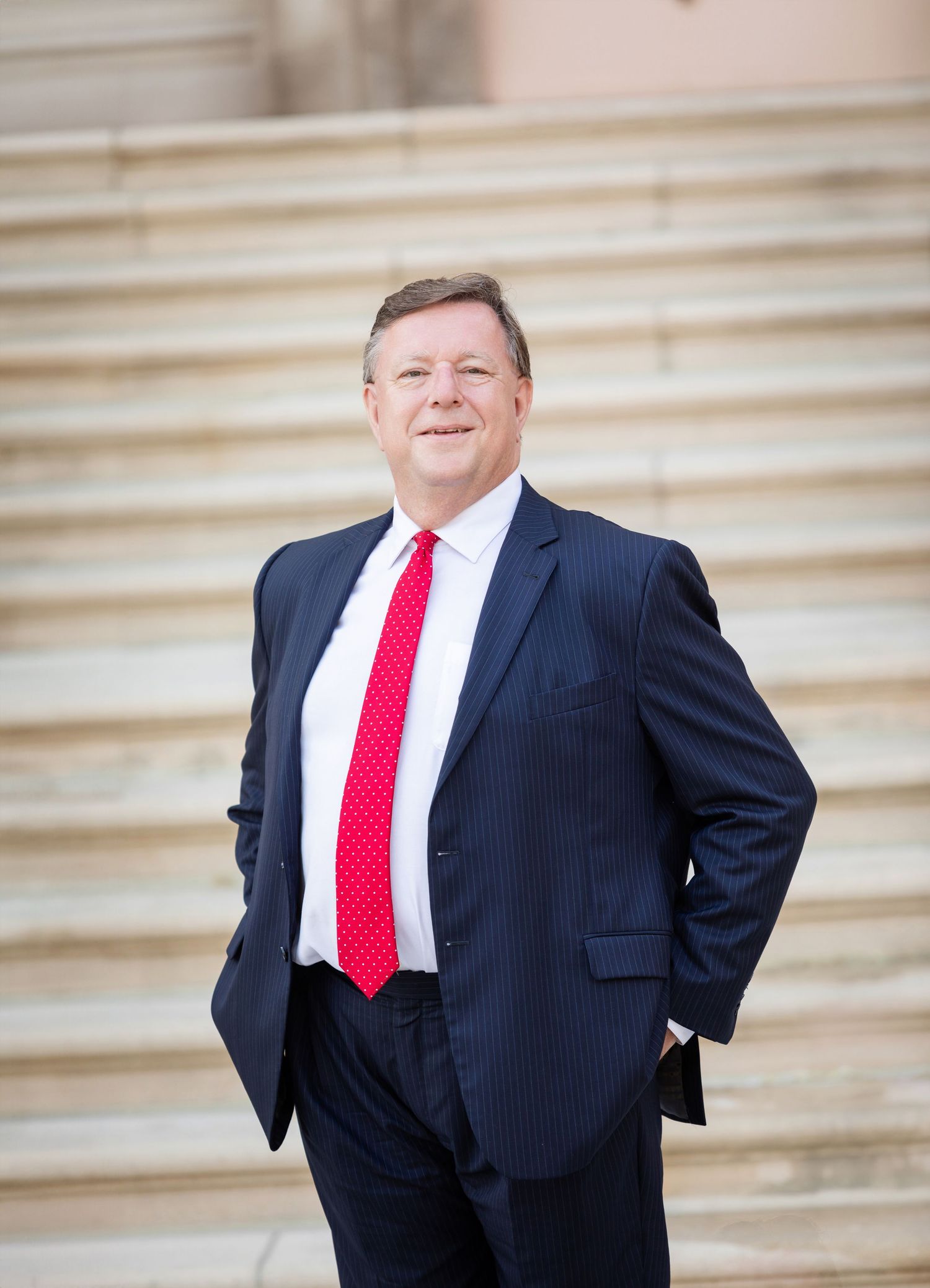 Professional man in navy pinstripe suit and red tie standing confidently on marble steps outdoors.