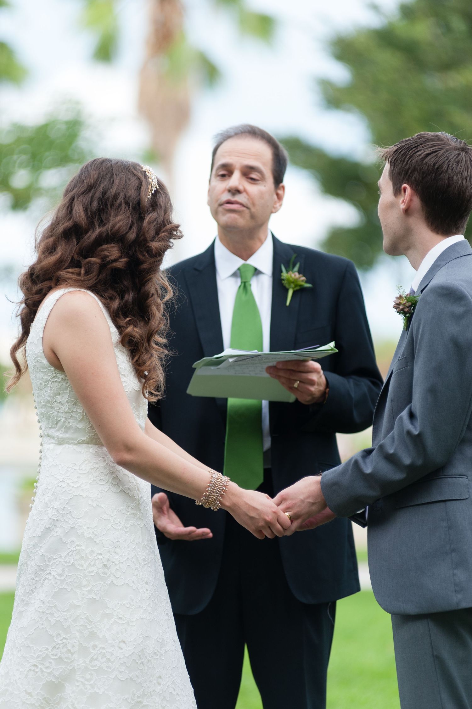 Bride and groom exchanging vows outdoors with officiant in green tie during wedding ceremony.