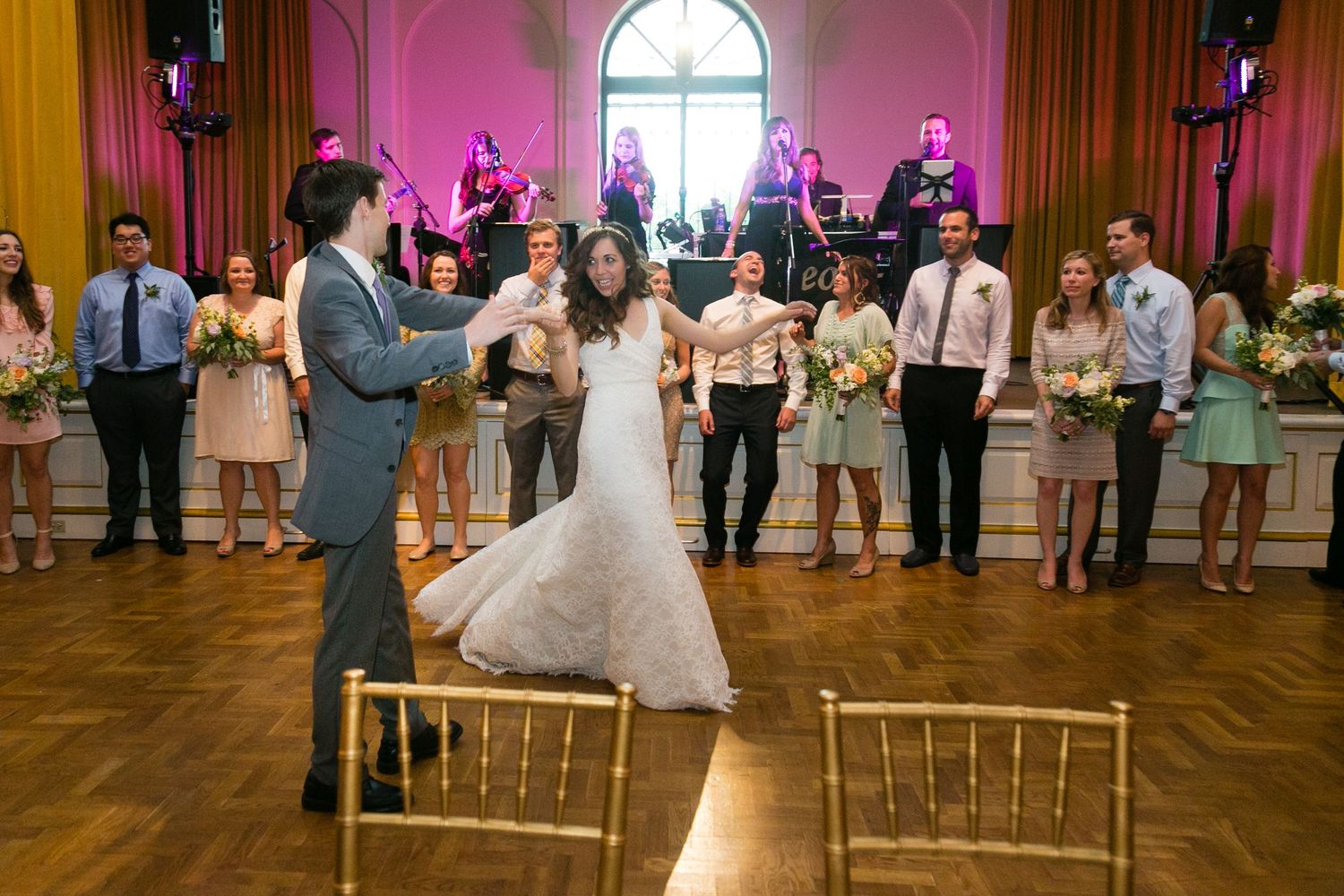 Bride and groom share first dance in elegant ballroom with live band and wedding guests watching.
