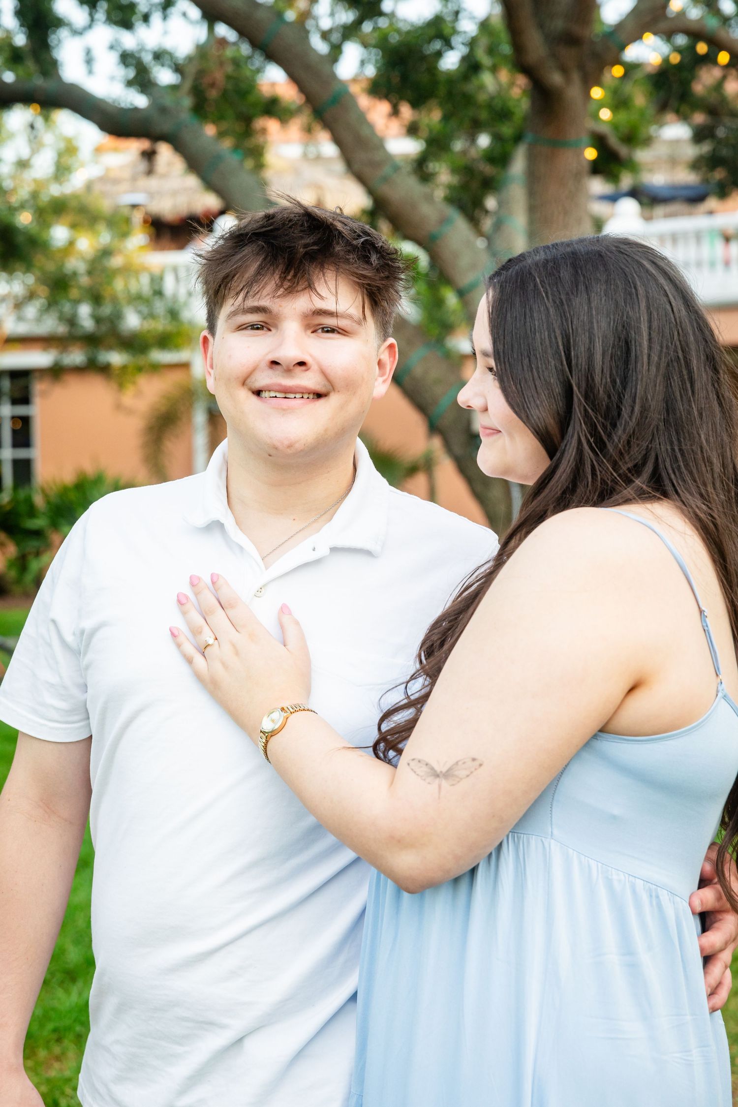 Young couple posing outdoors, man in white polo smiling as woman in blue dress places hand on his chest.