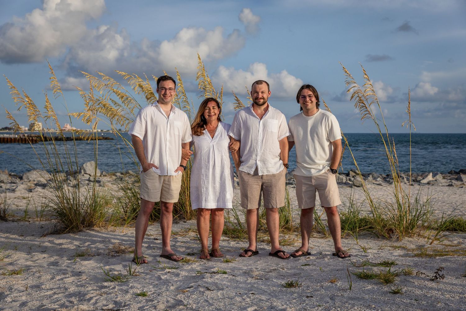 Family of four smiling together on a sandy beach with sea oats and blue ocean in the background.